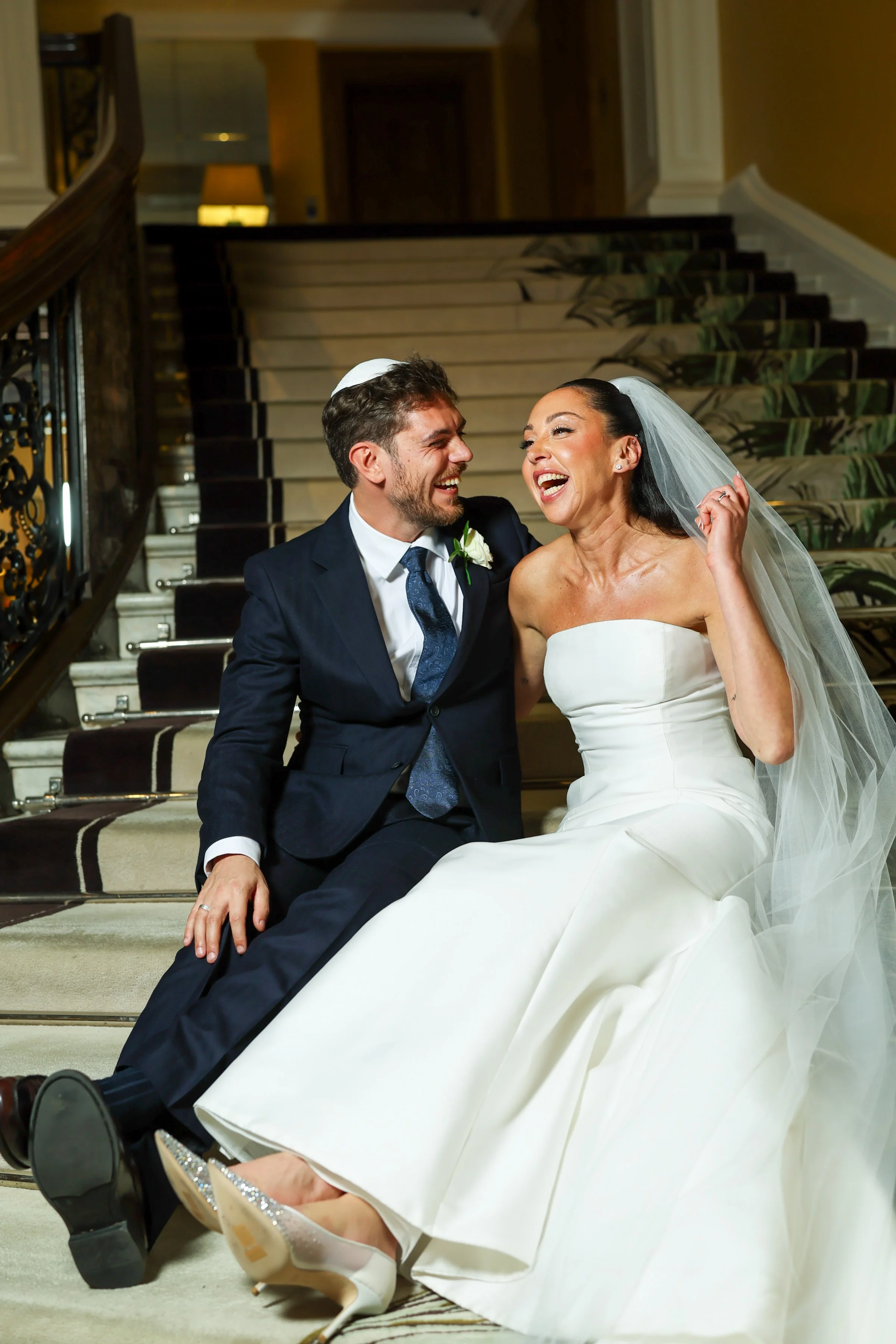 A newly married couple sitting on a staircase, smiling and laughing together. The groom is dressed in a dark suit and tie with a boutonniere, and the bride is wearing a strapless white wedding gown with a veil and sparkling shoes.