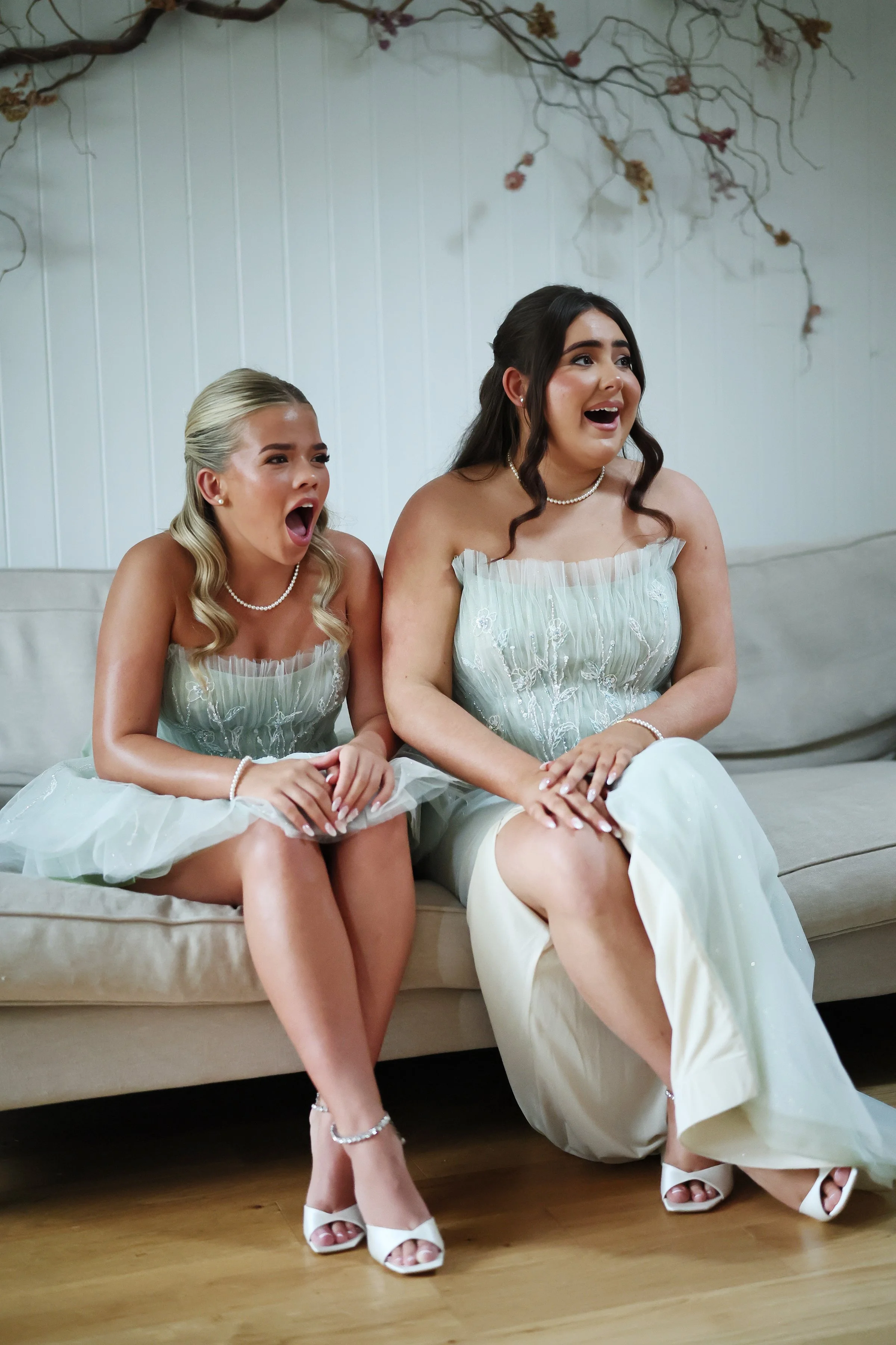 Two women wearing matching light-colored dresses with lace detail, pearl necklaces, and high heels, sitting on a beige couch with surprised or excited expressions.