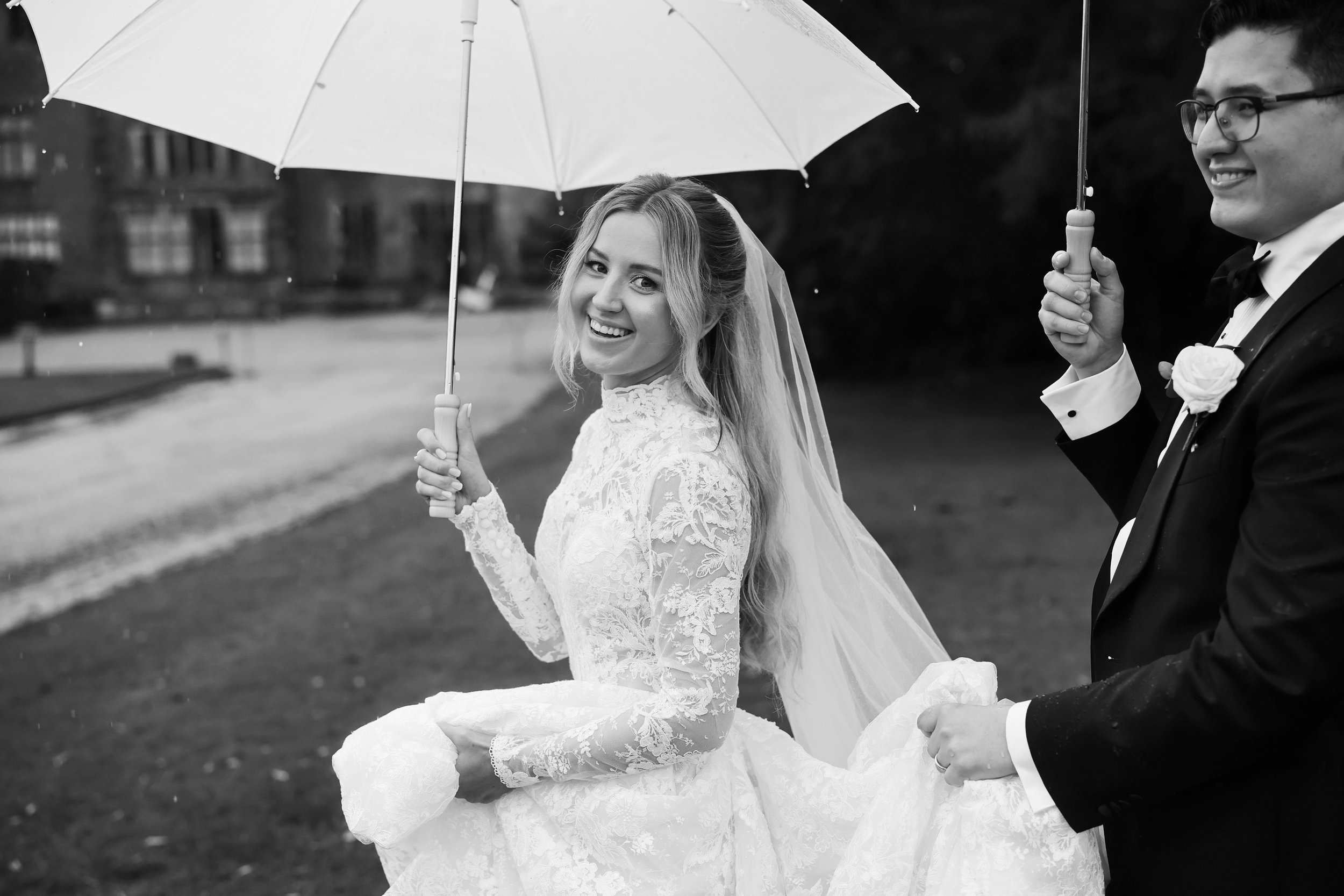 A smiling bride in a lace wedding dress holding a white umbrella, standing outdoors with a groom in a tuxedo holding a similar umbrella beside her.