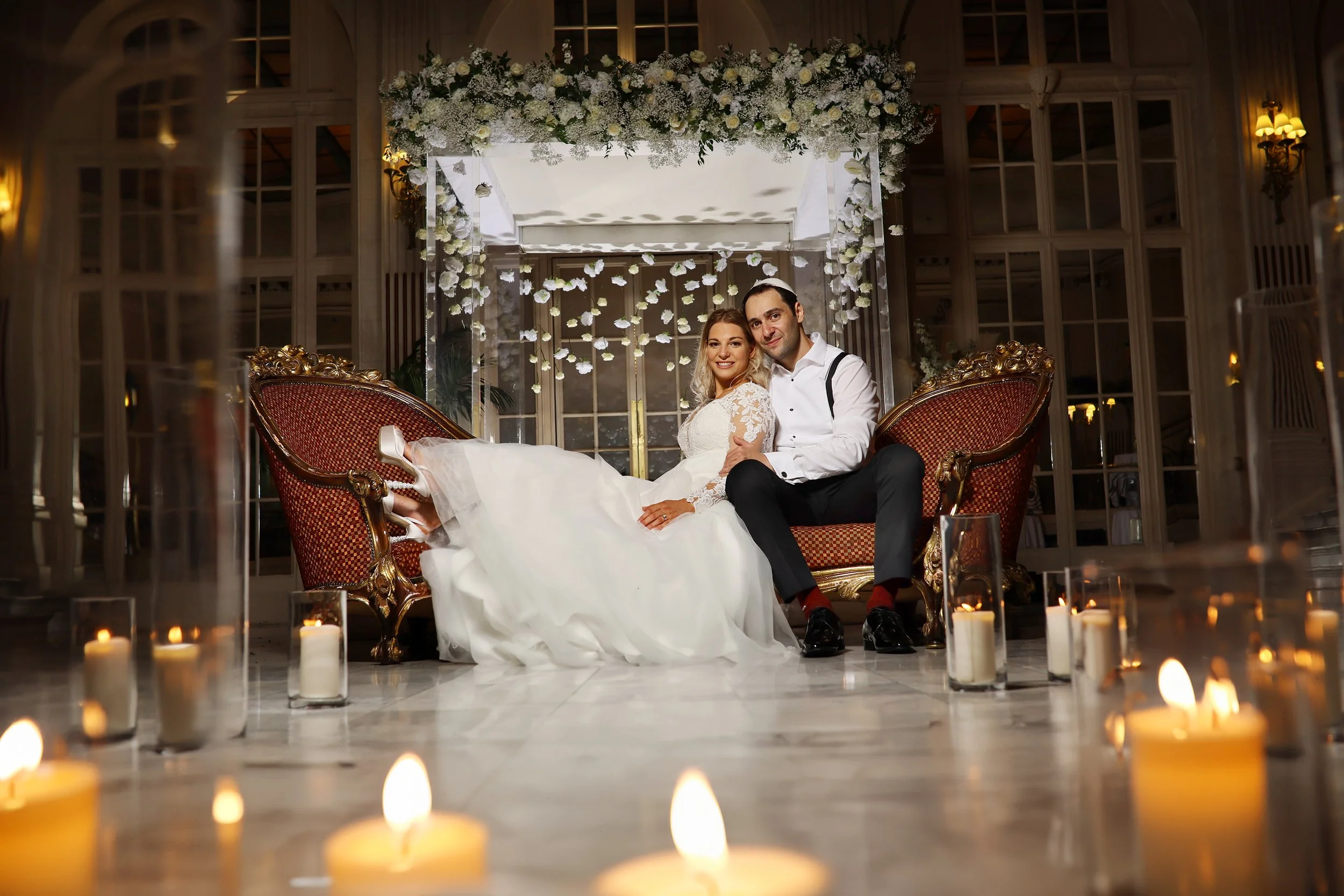 A newlywed couple sitting on a vintage-style red and gold sofa at their wedding reception, decorated with white flowers and candles.