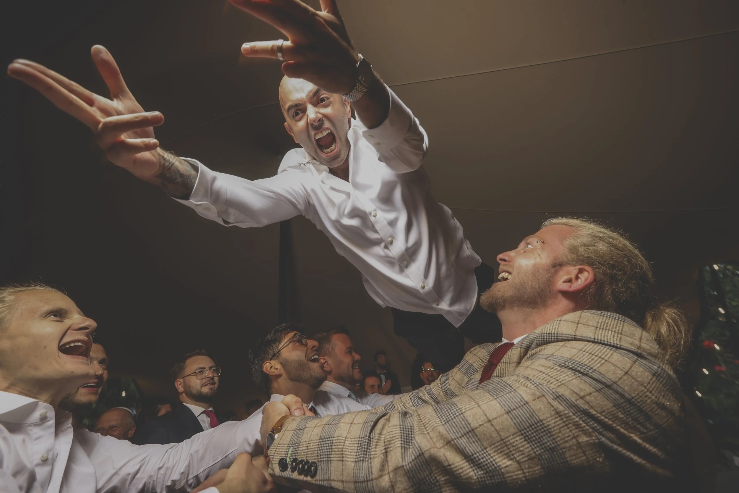 Group of people, including a man in a white shirt reaching out, sharing a joyful moment with a man in a patterned jacket in a crowded indoor setting.