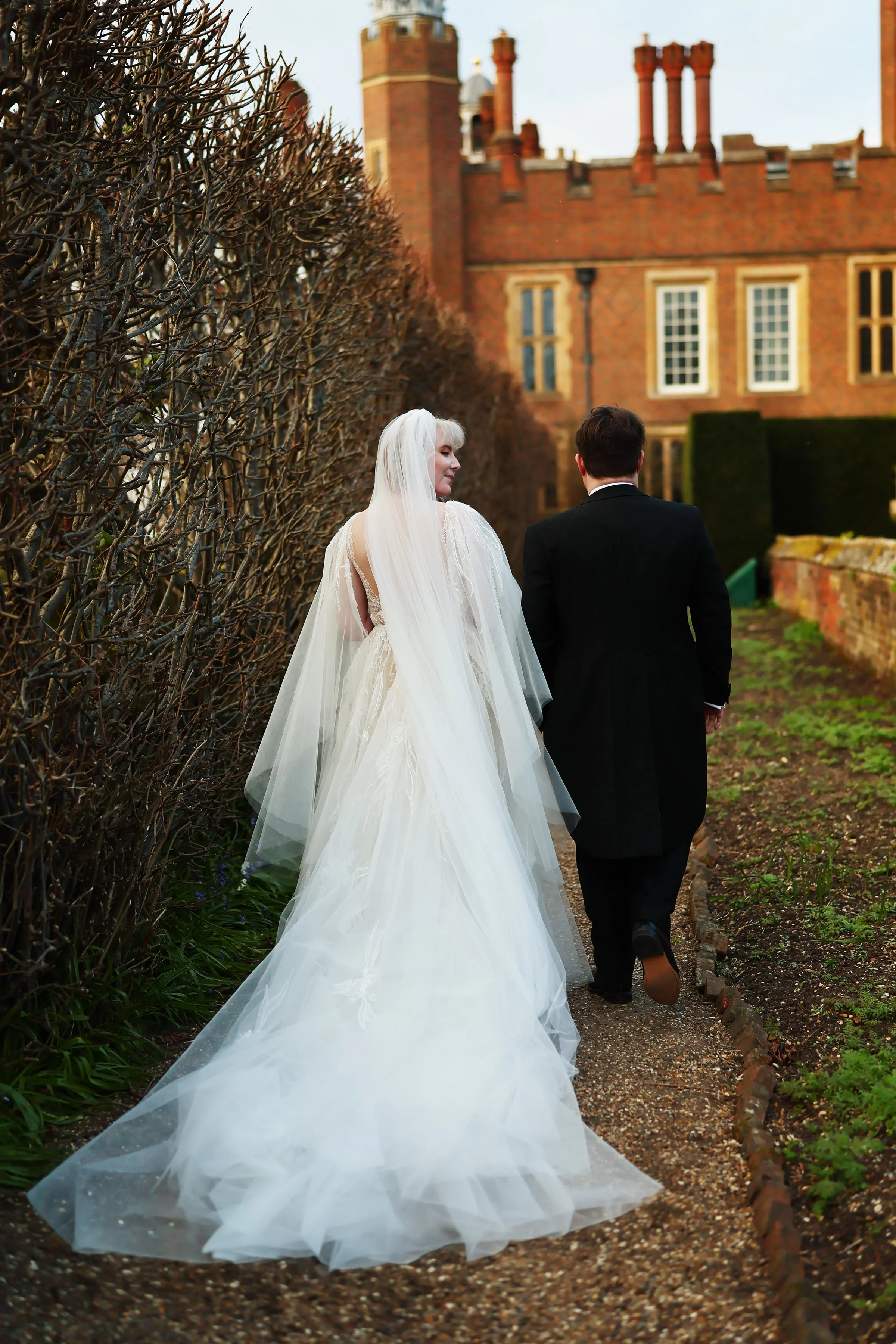 A bride and groom walking away hand in hand on a garden path outside a brick building with tall windows and chimneys, the bride wearing a white gown and veil, and the groom in a black suit.