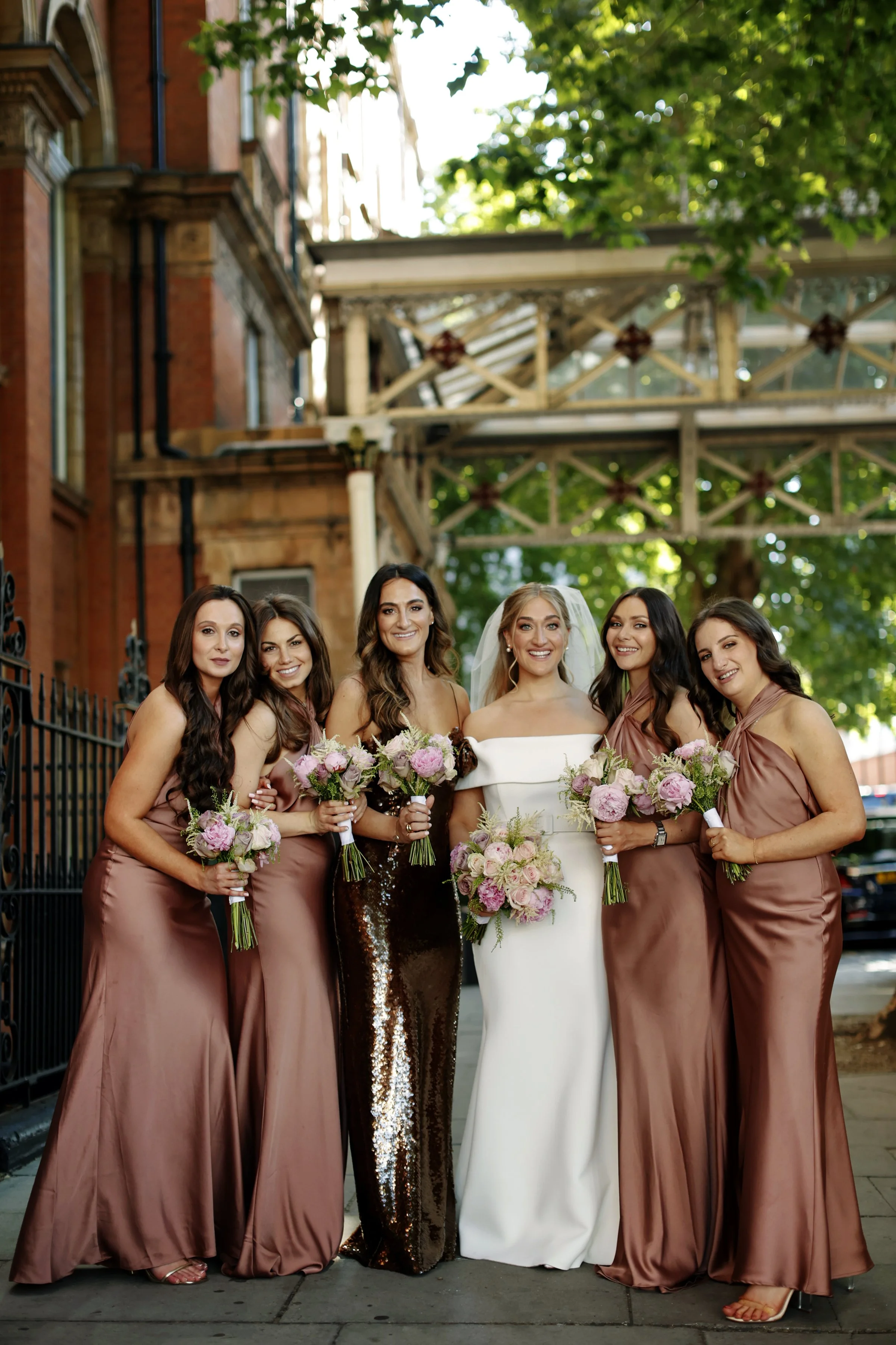 Group of six women, one in a white wedding dress, five in matching mauve dresses, holding bouquets, outdoors in front of a brick building.