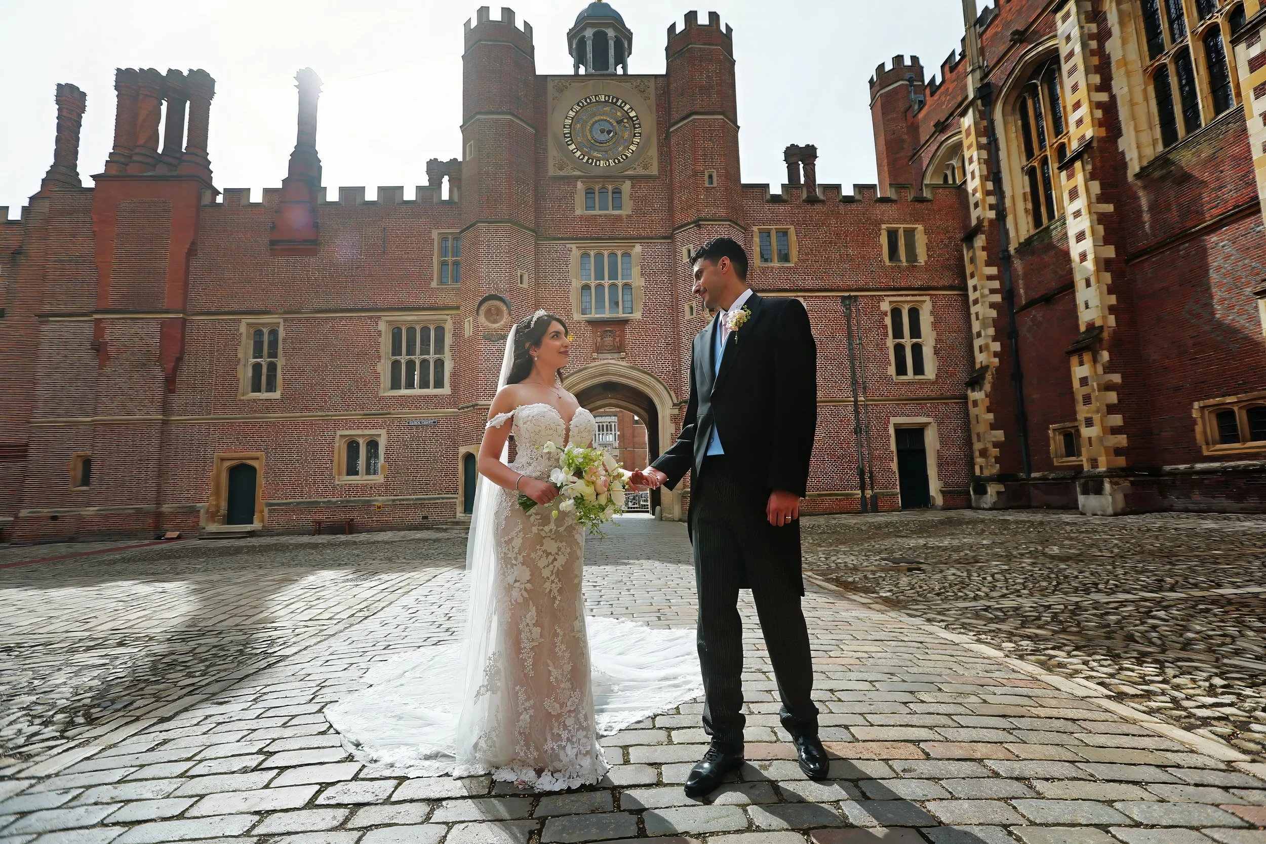 A bride and groom holding hands and facing each other in front of a historic brick castle-like building, with the bride in a lace wedding gown and the groom in a black tuxedo. The bride is holding a bouquet of flowers. The scene is lit by sunlight.