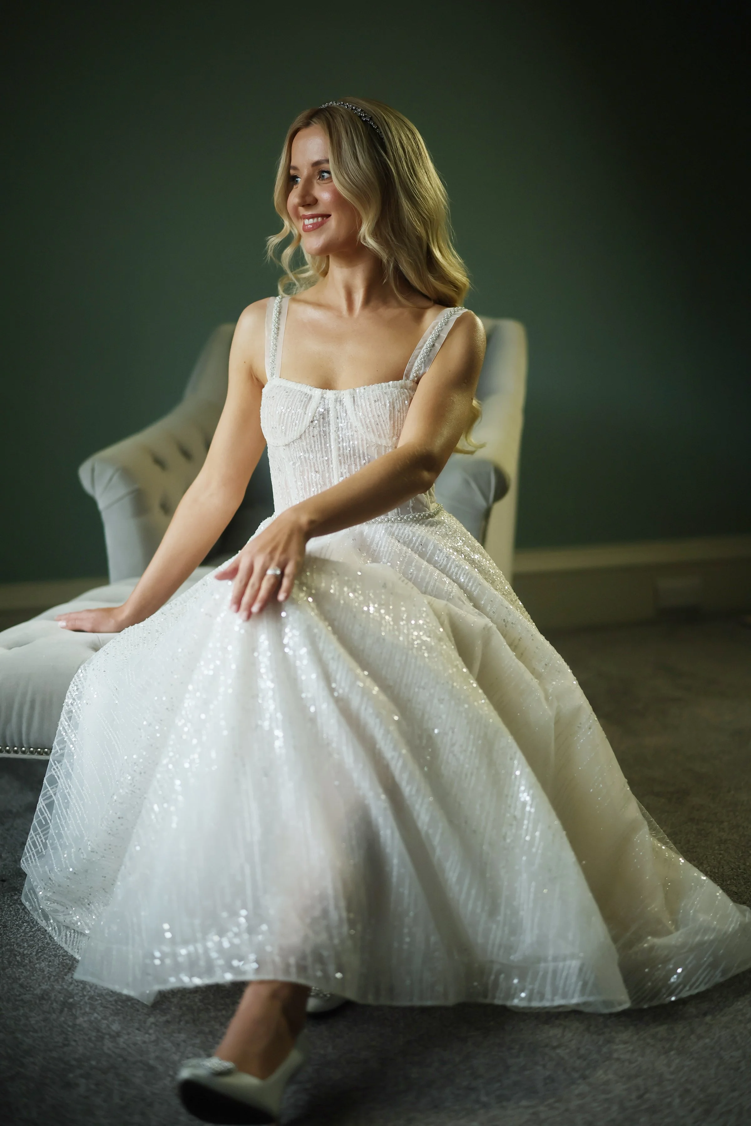A young woman with blonde hair in loose waves, wearing a sparkly white dress with spaghetti straps and a headband, sitting on a beige tufted armchair with a dark green wall behind her.
