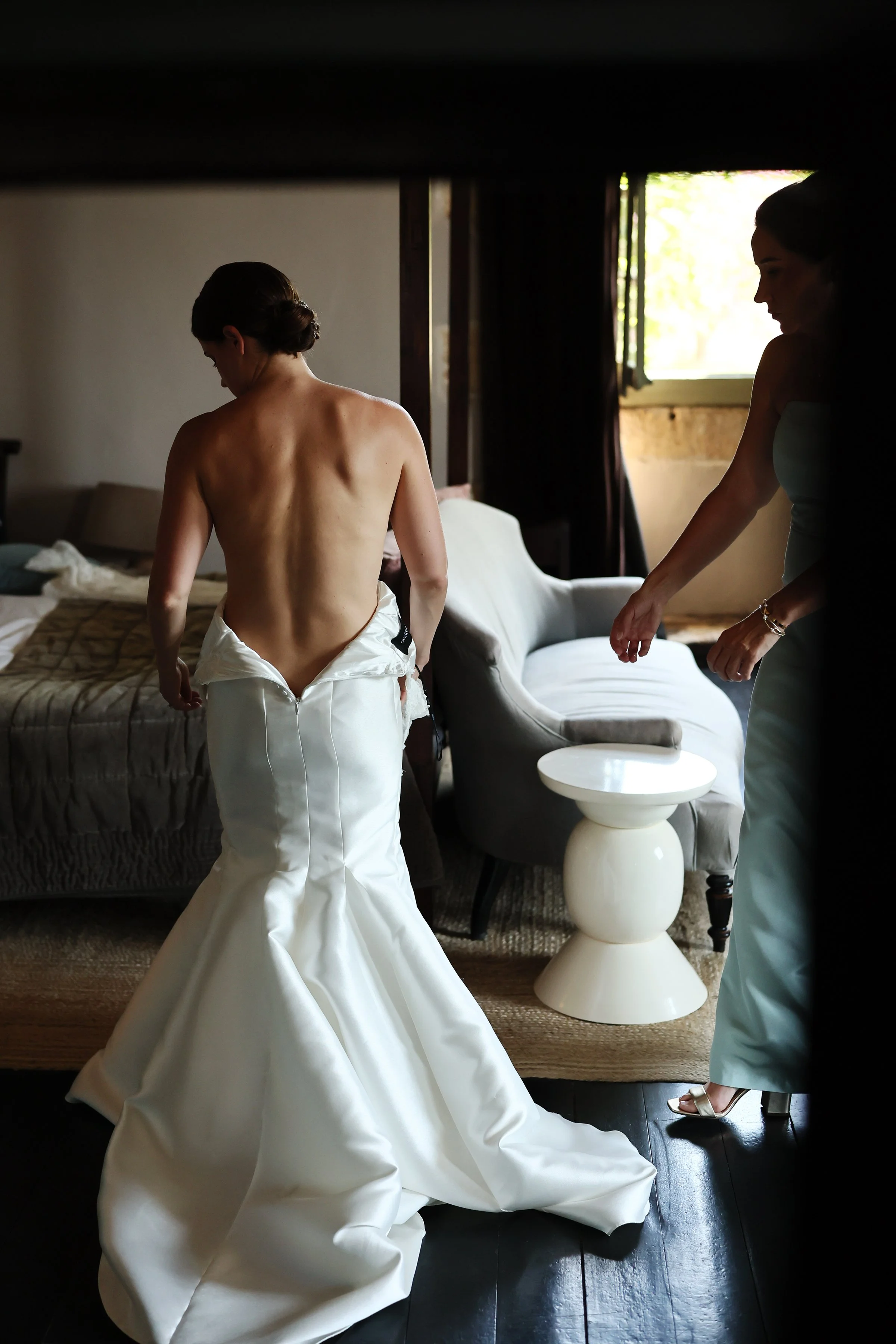 A woman getting dressed in a wedding gown, with another woman assisting her, in a softly lit room with a window and furniture.
