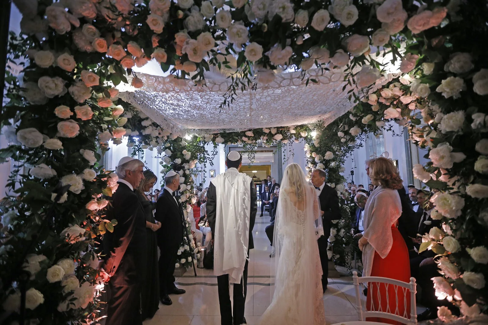 A wedding ceremony indoors with the bride and groom standing under a floral arch. The arch is decorated with white roses and greenery. Guests are seated and standing around, watching the ceremony.