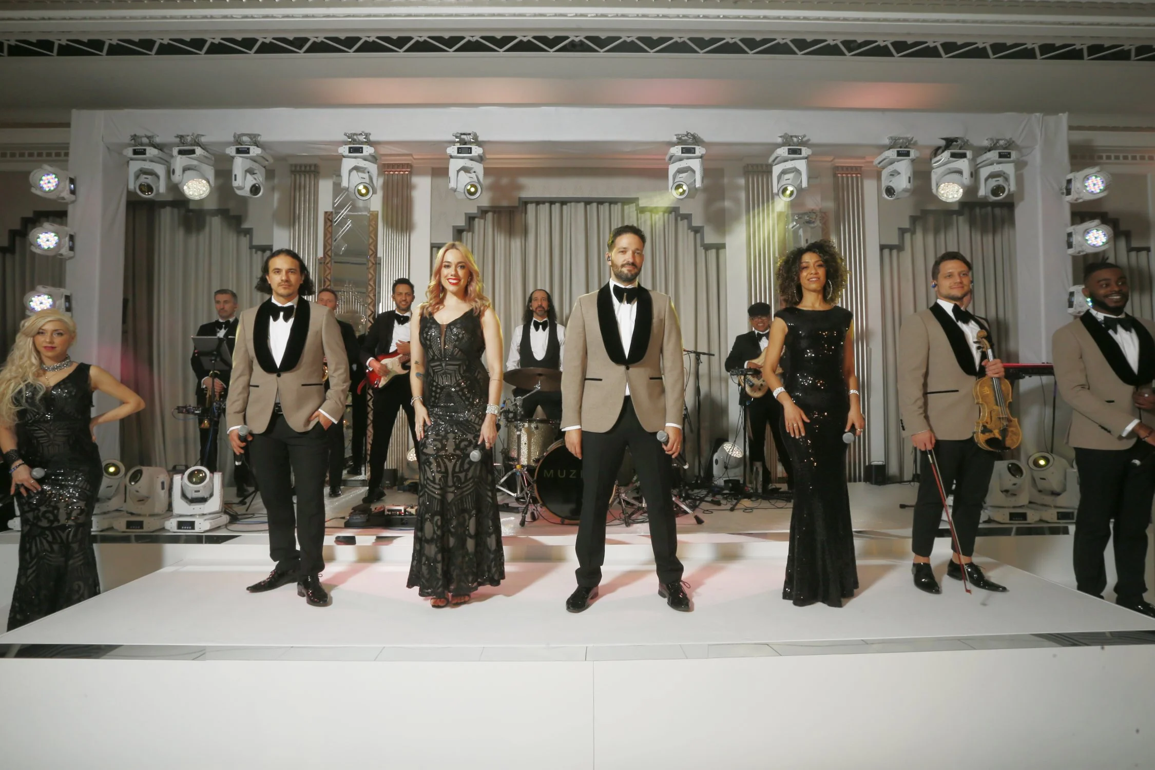 A group of performers on stage, dressed in tuxedos and black dresses, posing in front of a band with musical instruments and bright stage lighting.