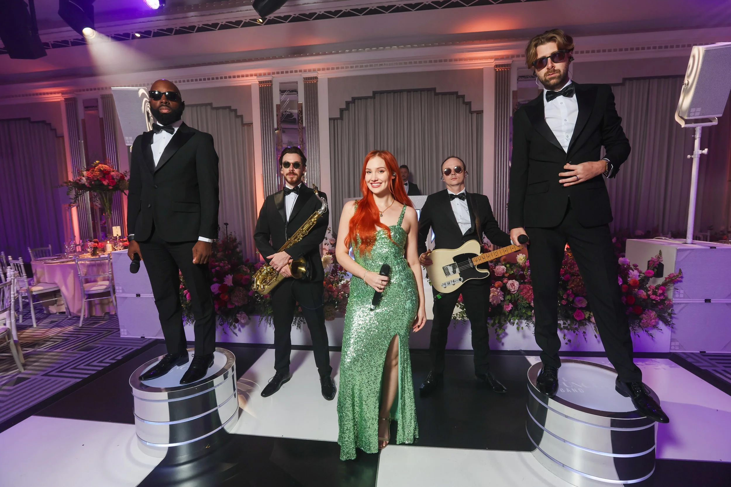 A group of wax statues of musicians posing on a stage, with a woman in a green dress in the center holding a microphone, and four men in tuxedos with instruments and sunglasses around her, in a decorated event hall with floral arrangements.