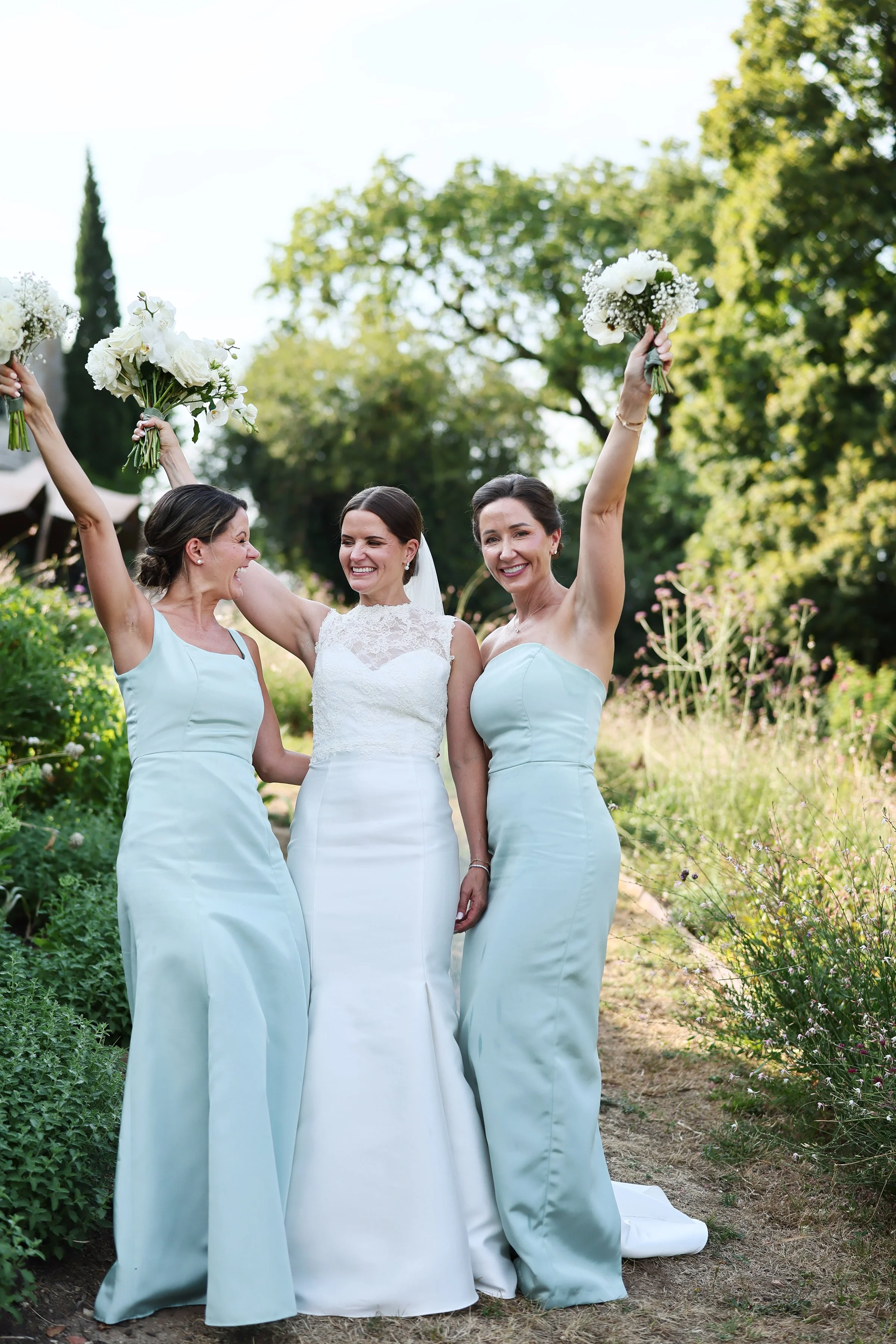 Three women in wedding and bridesmaid dresses celebrating outdoors, holding bouquets, smiling, with trees in the background.