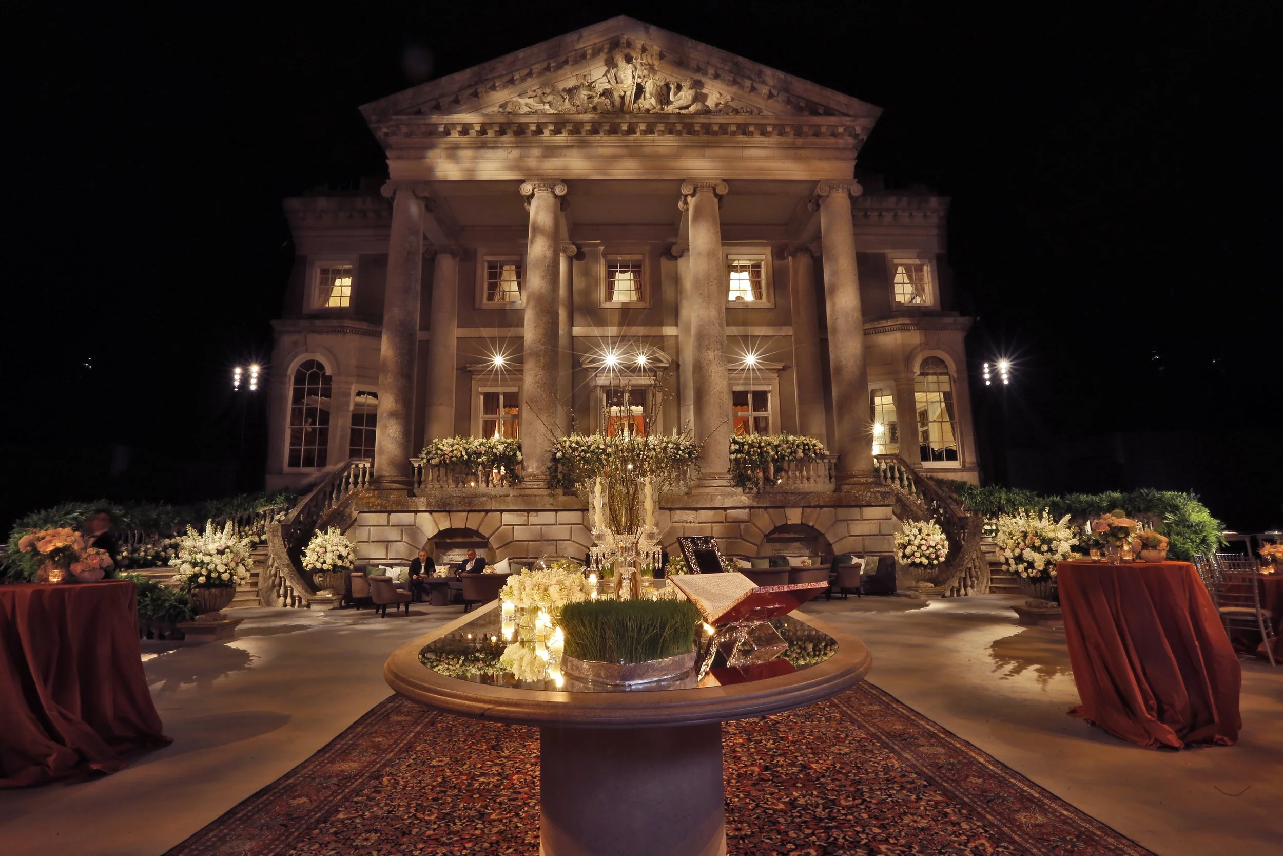 Night view of a grand, illuminated mansion with classical architecture, large columns, and a wide staircase, decorated with flowers and elegant furnishings for an event.