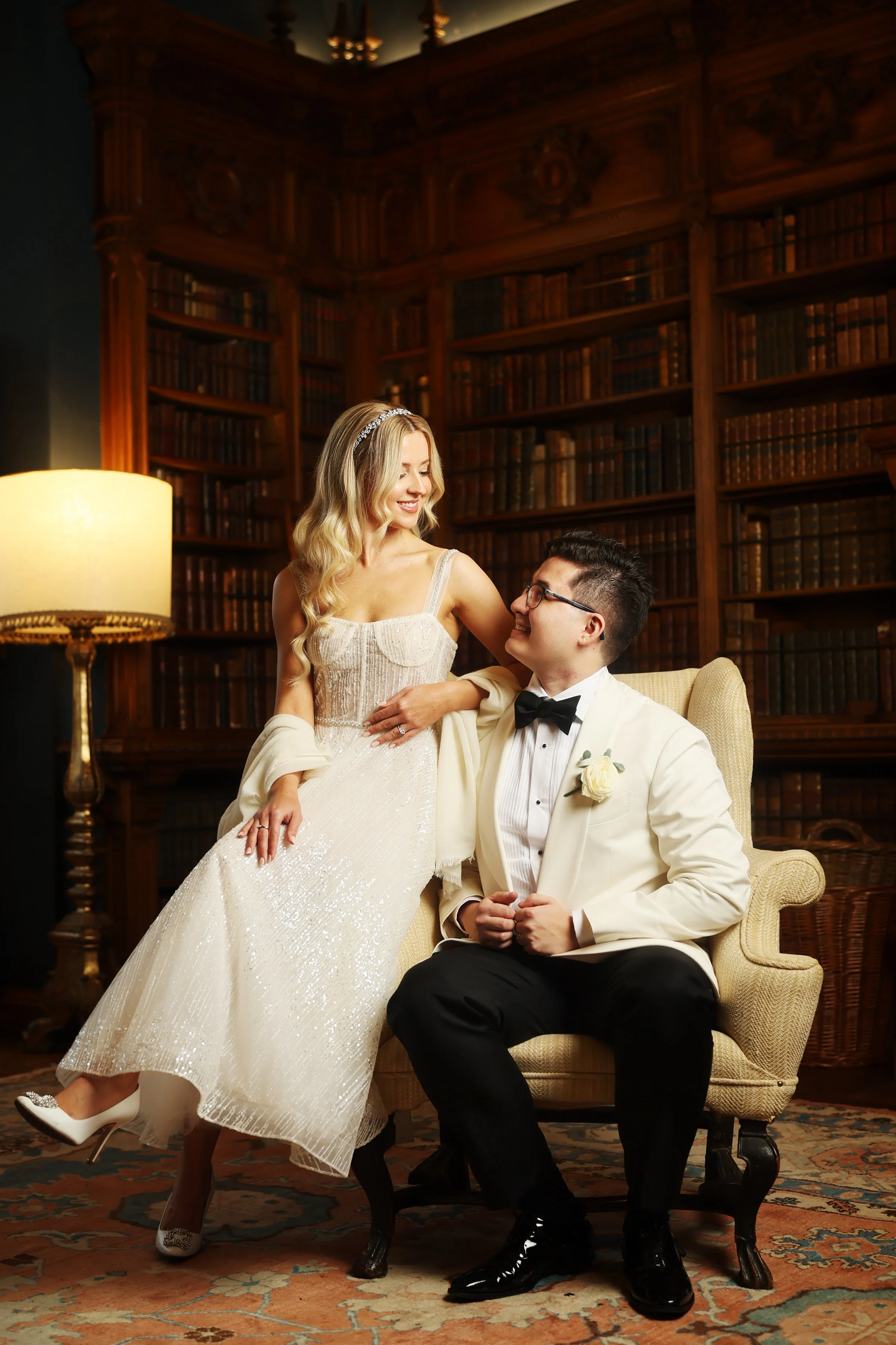A woman in a wedding dress and a man in a tuxedo are sitting in a library. The woman is smiling at the man, who is looking up at her. The library has tall wooden bookshelves filled with books and a warm lamp beside them.