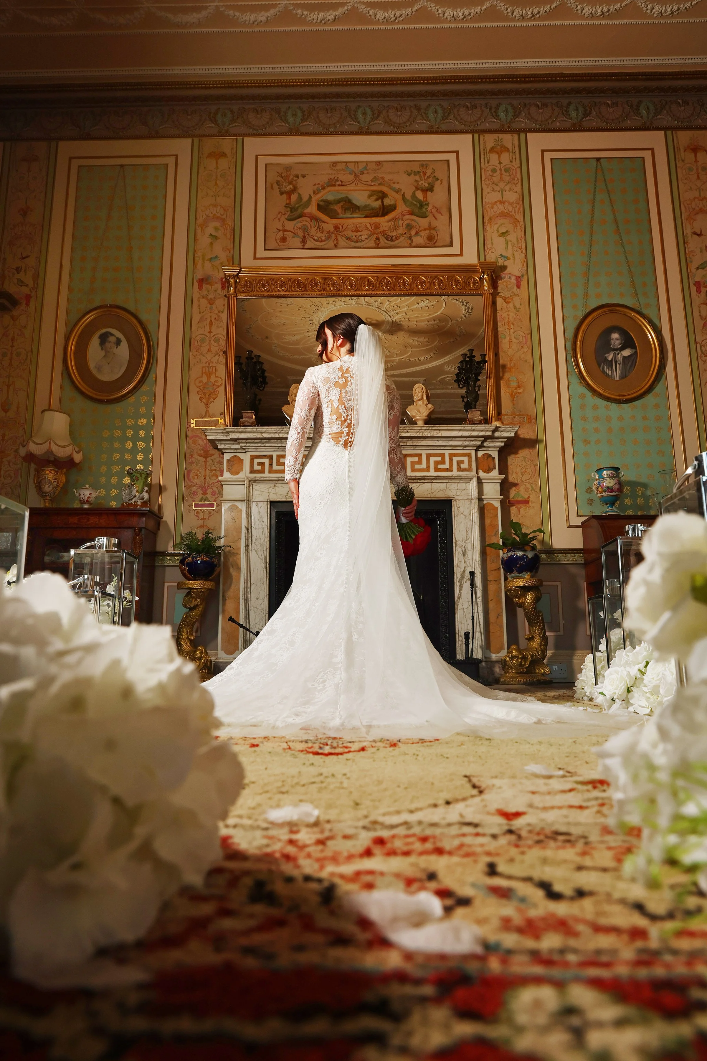 A bride standing in front of a fireplace in an ornate room, wearing a white lace wedding dress and veil. The room is decorated with framed portraits, vases, and intricate wall designs, with flower petals scattered on the floor.