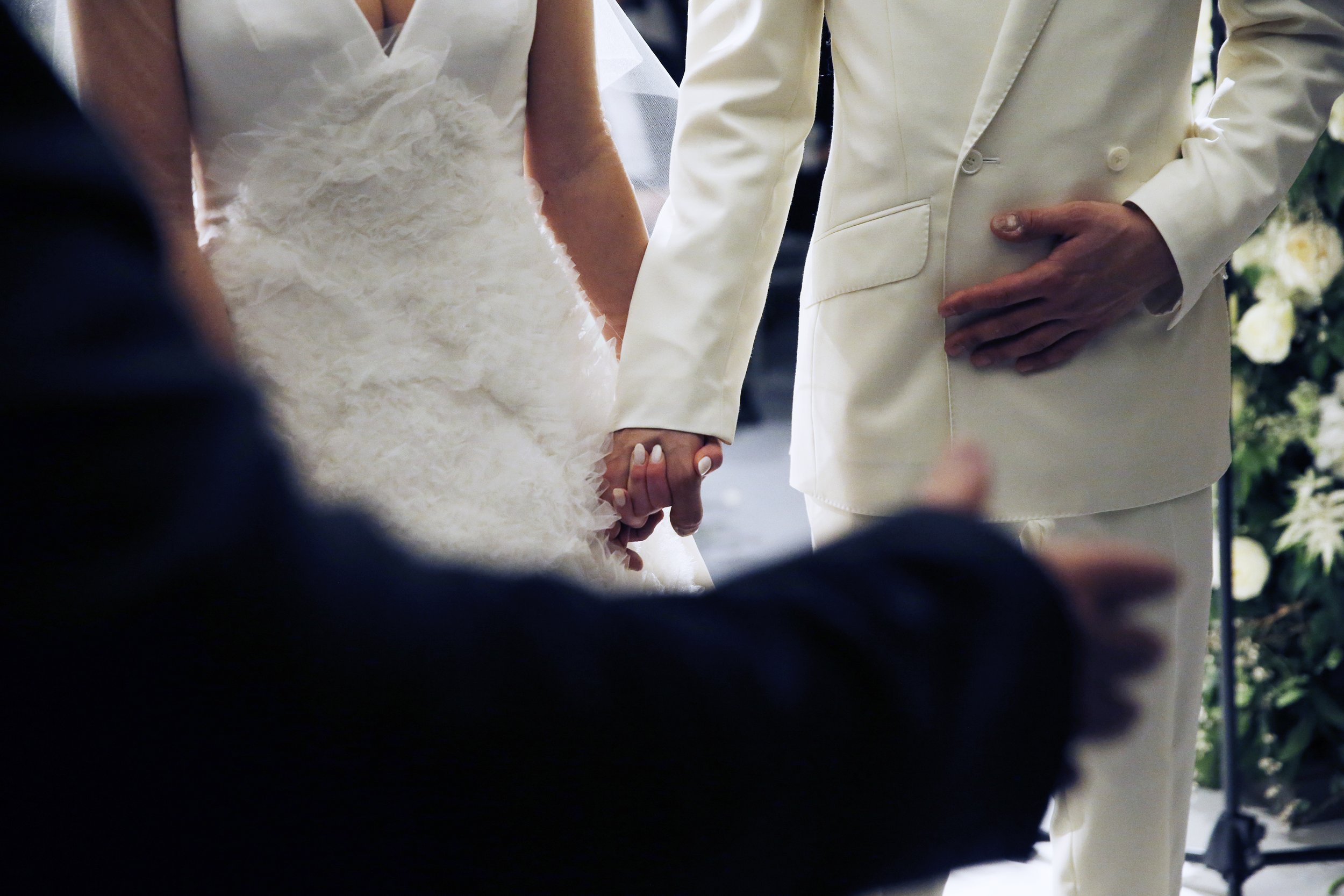 A couple holding hands during their wedding ceremony, with the bride in a white wedding gown and the groom in a cream-colored suit.