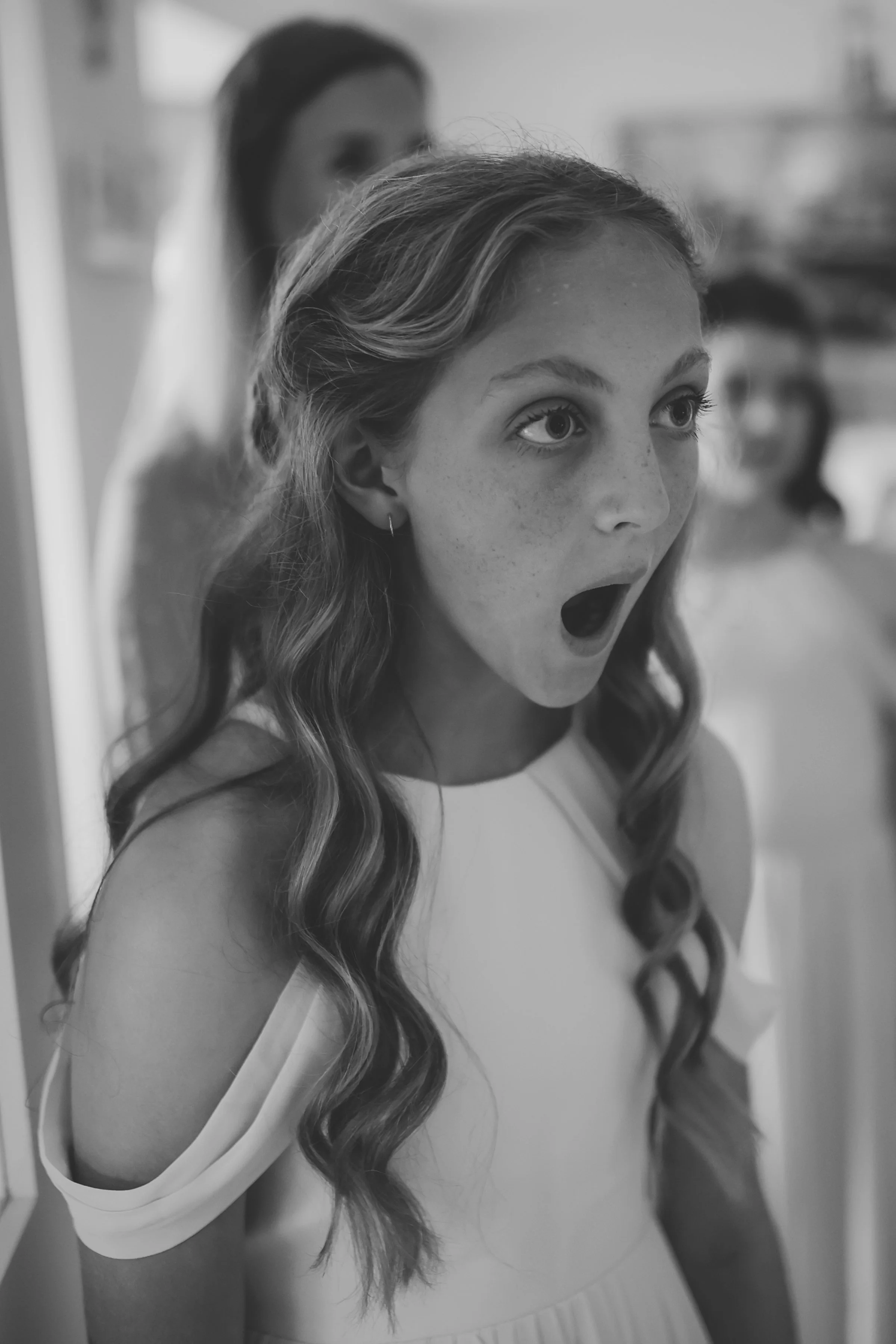 A young girl with long, wavy hair and freckles, wearing a sleeveless top and shorts, with her mouth open as if surprised or excited. Another girl with dark hair is blurred in the background.