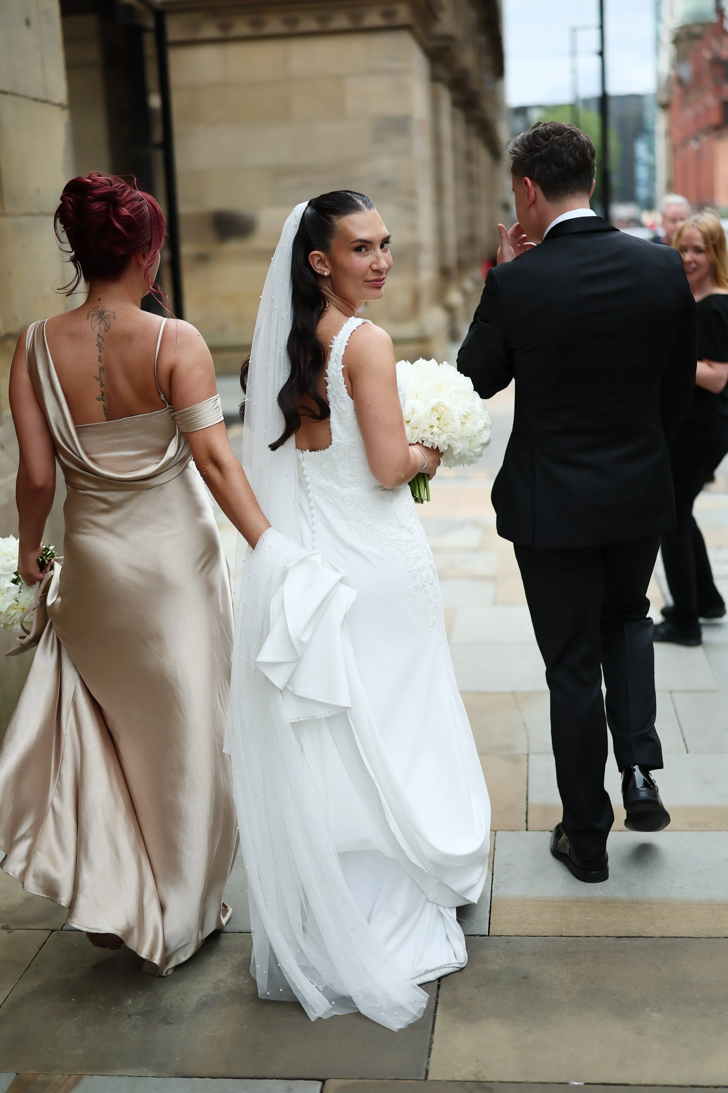 A bride in a white wedding dress holding a bouquet, walking with a woman in a champagne satin gown and a man in a black tuxedo outside a historic building with stone walls, with other people in the background.