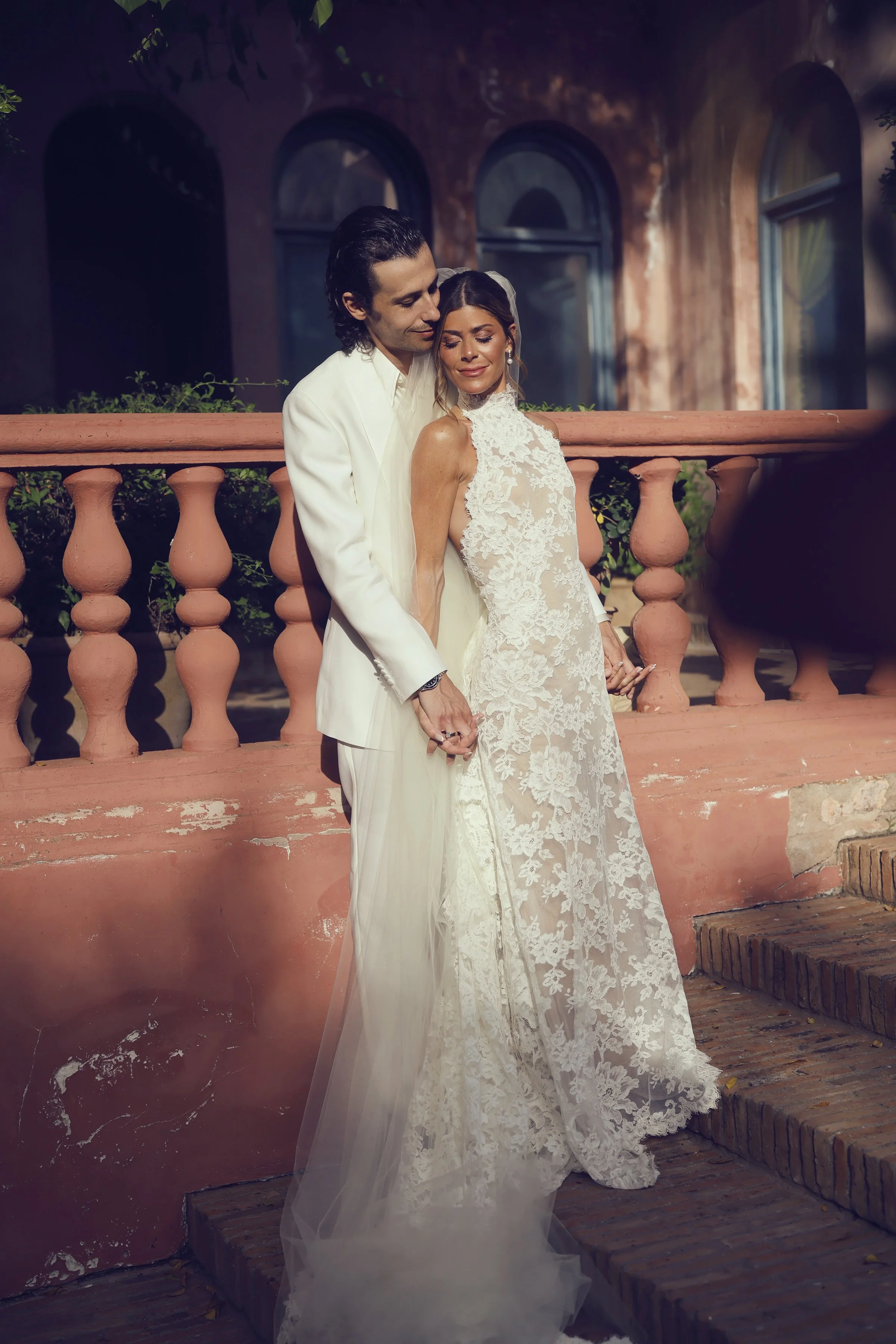 A couple in wedding attire standing on brick stairs outdoors, holding hands, with the man in a white suit and the woman in a lace wedding dress, embracing each other tenderly.