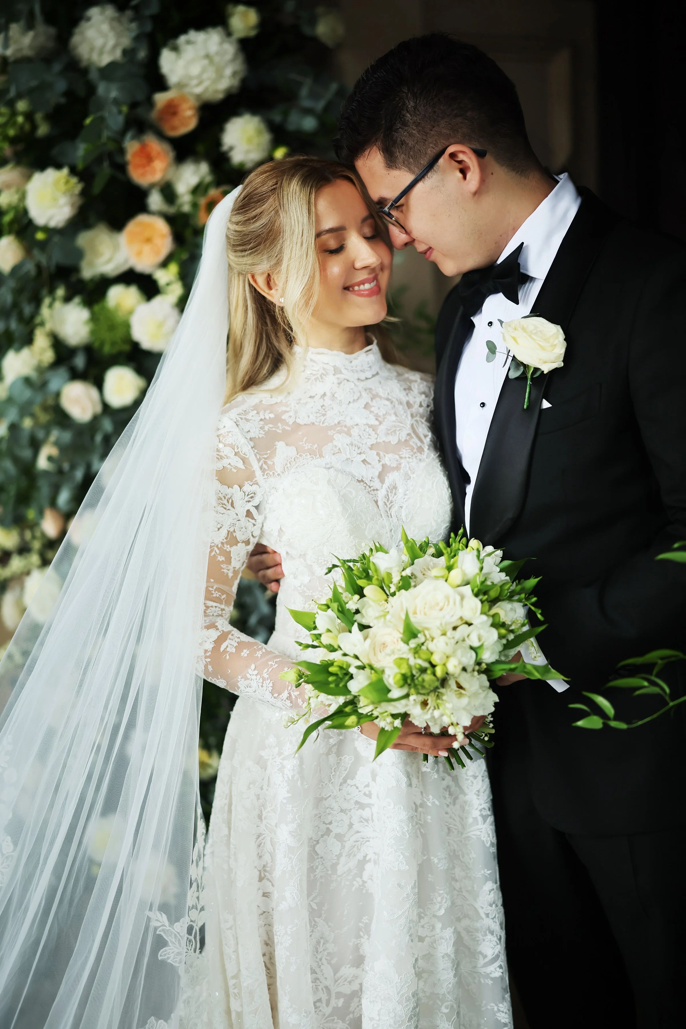 A bride and groom with their foreheads touching, smiling, during their wedding ceremony. The bride is wearing a lace wedding gown, and the groom is in a black tuxedo with a white boutonniere. The bride is holding a bouquet of white and cream flowers.