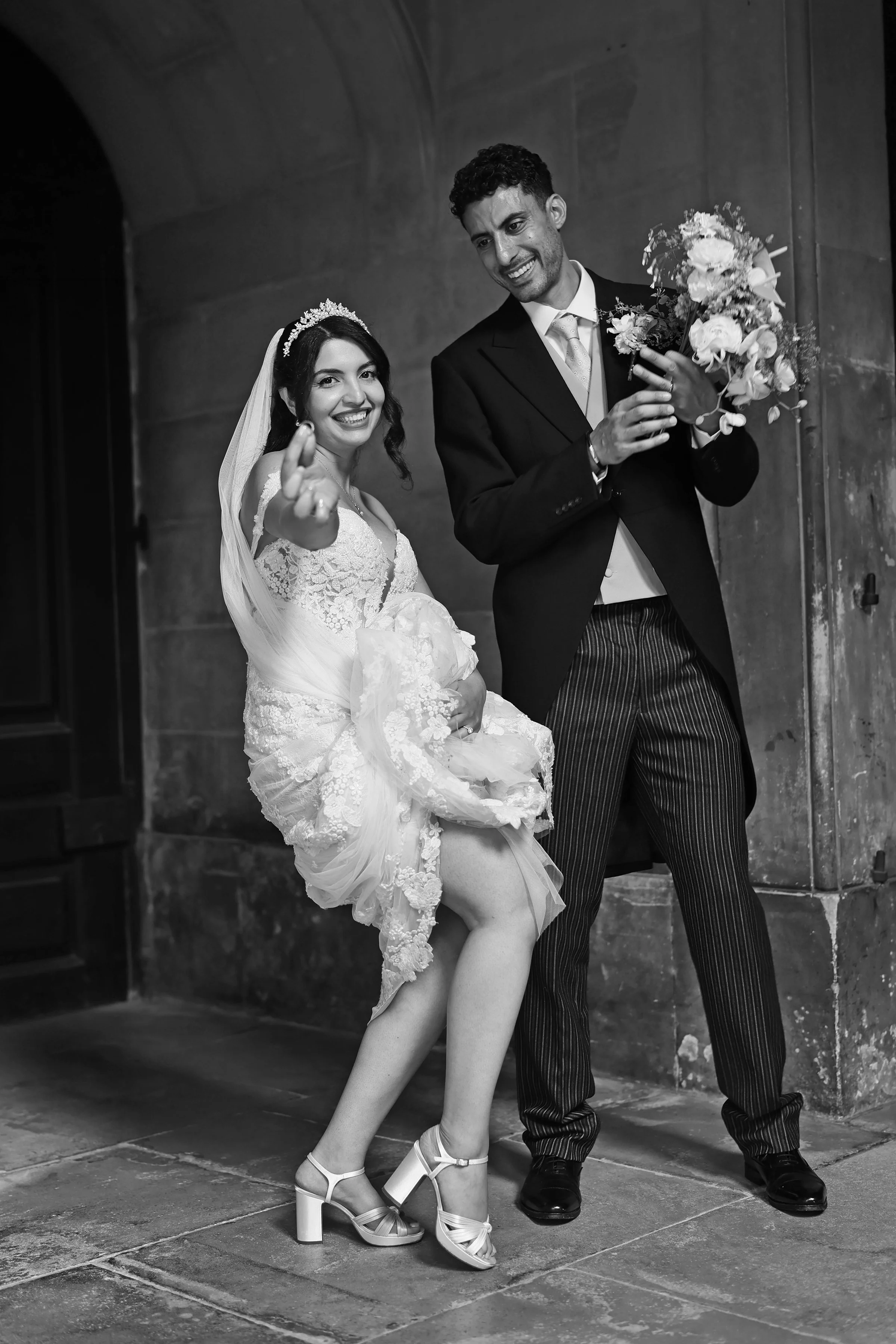 Black and white photo of a bride and groom standing indoors. The bride is wearing a lace wedding dress, veil, and high heels, smiling and pointing towards the camera. The groom is dressed in a suit, holding a bouquet of flowers, and smiling.