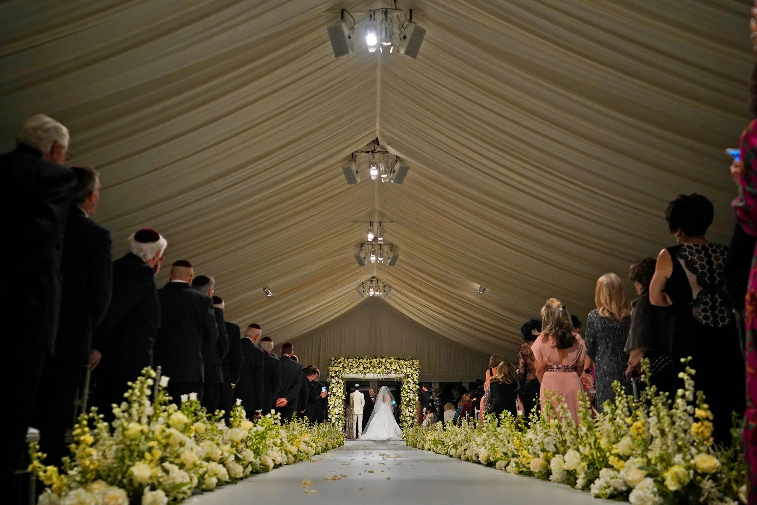 Wedding ceremony inside a large decorated tent with floral arrangements along the aisle. The bride and groom are standing at the altar, facing away from the camera, surrounded by guests.