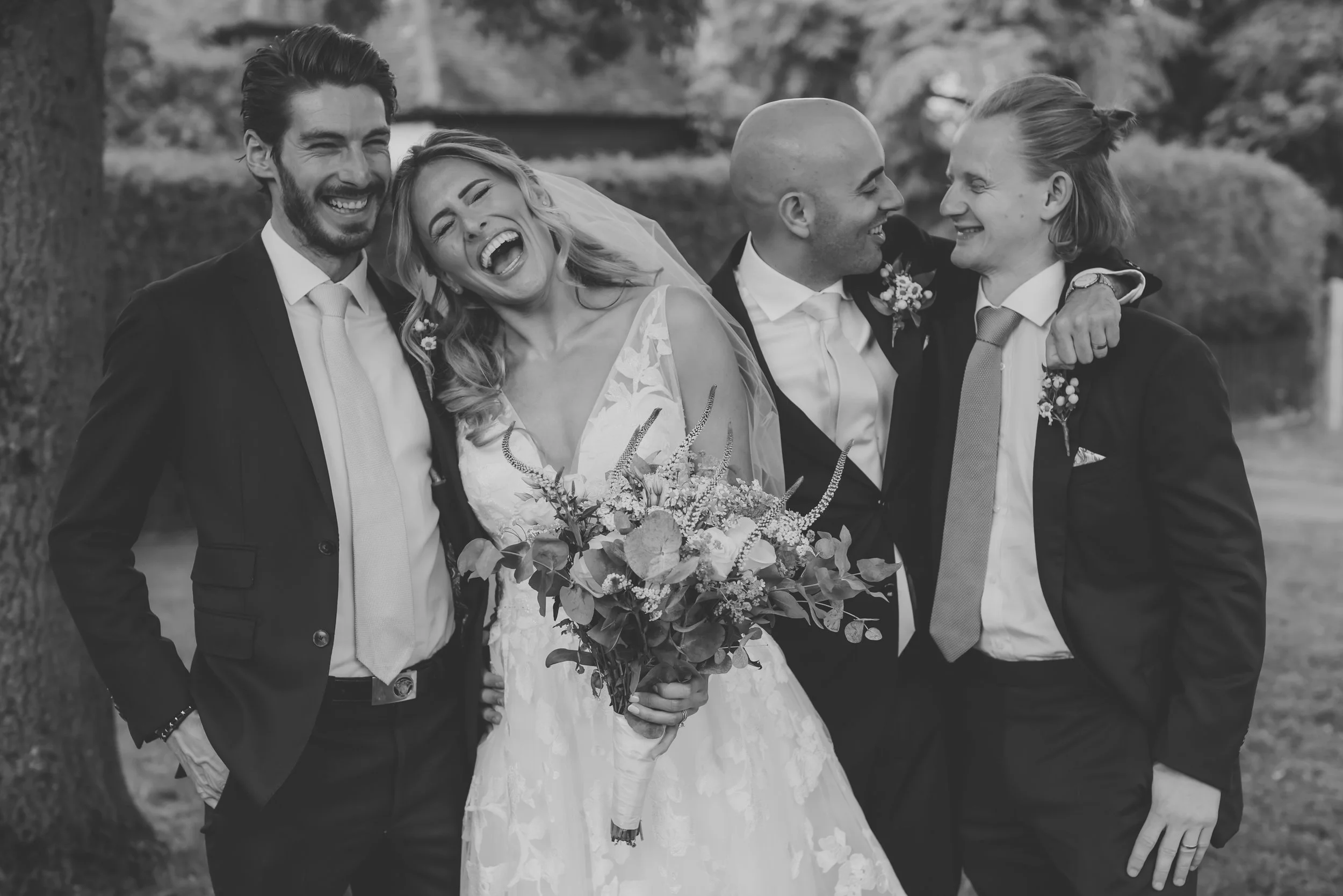 Black and white photo of five friends or family members smiling and laughing outdoors at a wedding, with the bride in the center holding a bouquet, and the others standing close around her with arm around each other.