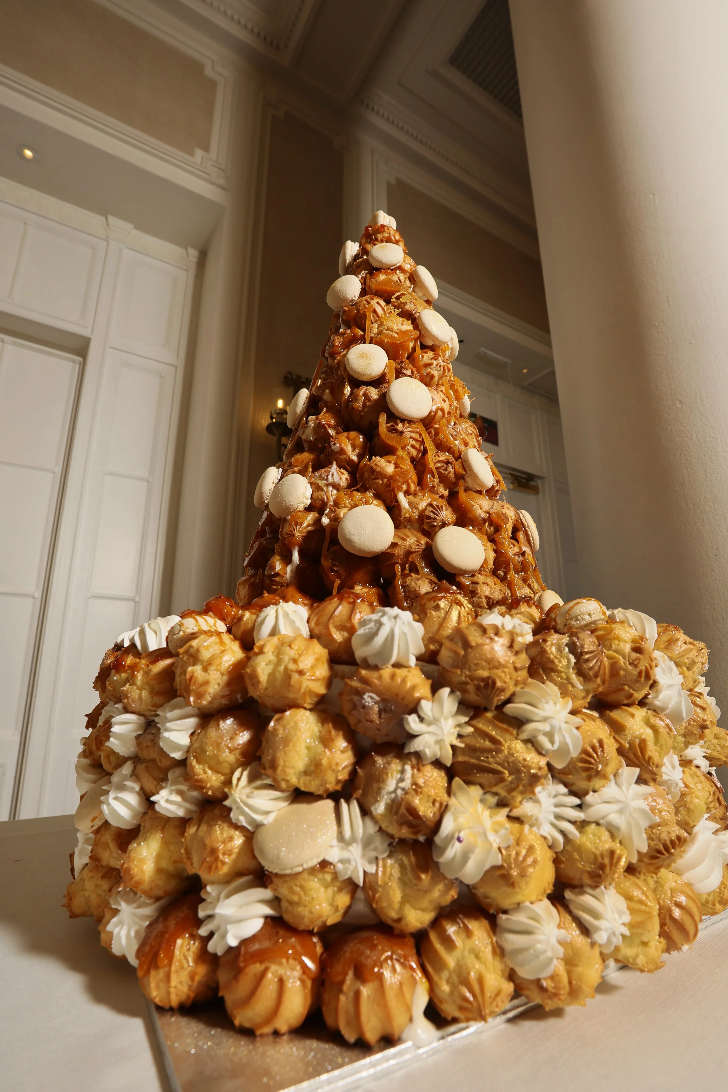 A layered croquembouche dessert tower decorated with caramel popcorn, white meringue kisses, and caramel candies inside a restaurant or banquet hall.