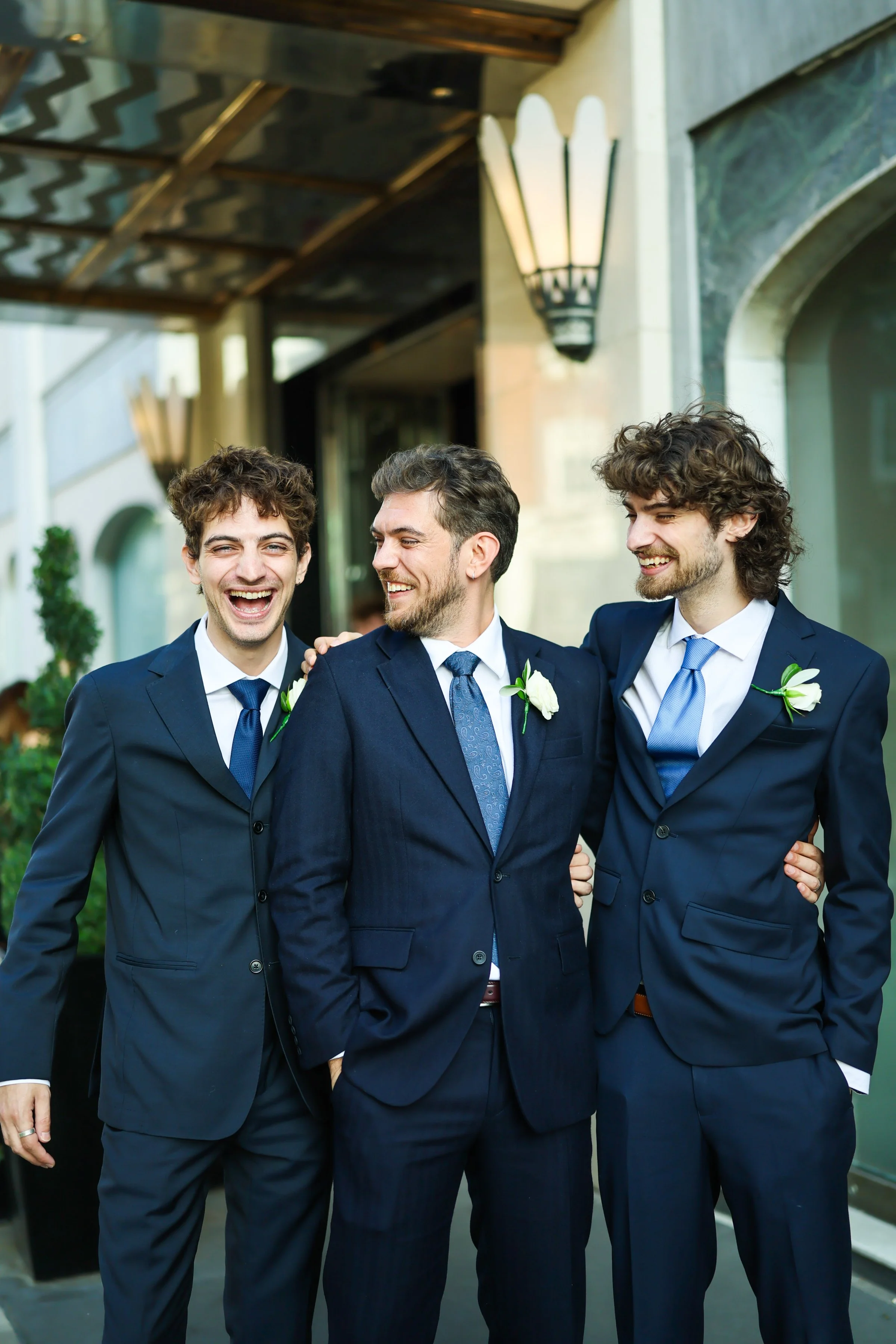 Three men in suits smiling and talking to each other outdoors near a building with a stone archway and lanterns.