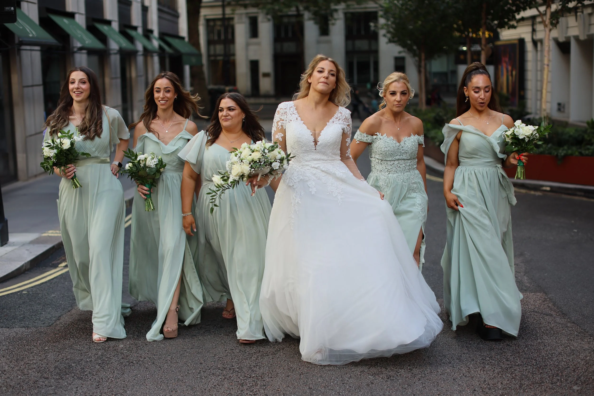 A bride in a white wedding gown walking with six bridesmaids on a city street, all holding bouquets of white flowers.