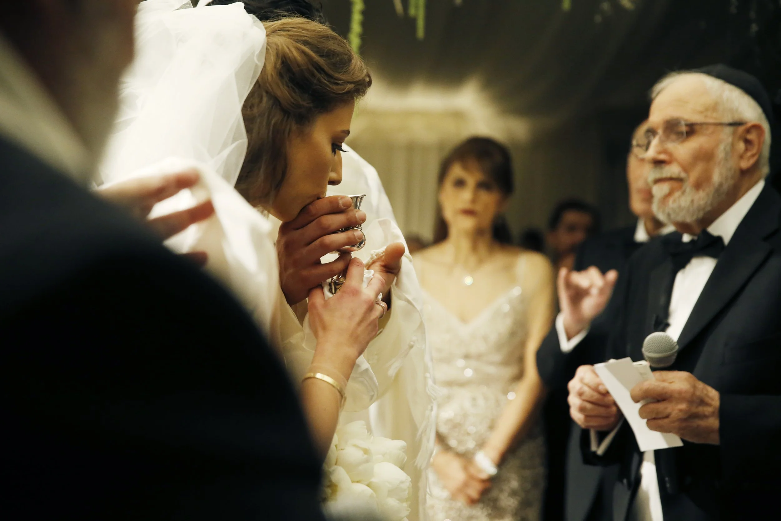 A woman dressed as a bride holding a silver cup and appearing to kiss it, while a man in a tuxedo with a microphone reads from a paper at a formal event.
