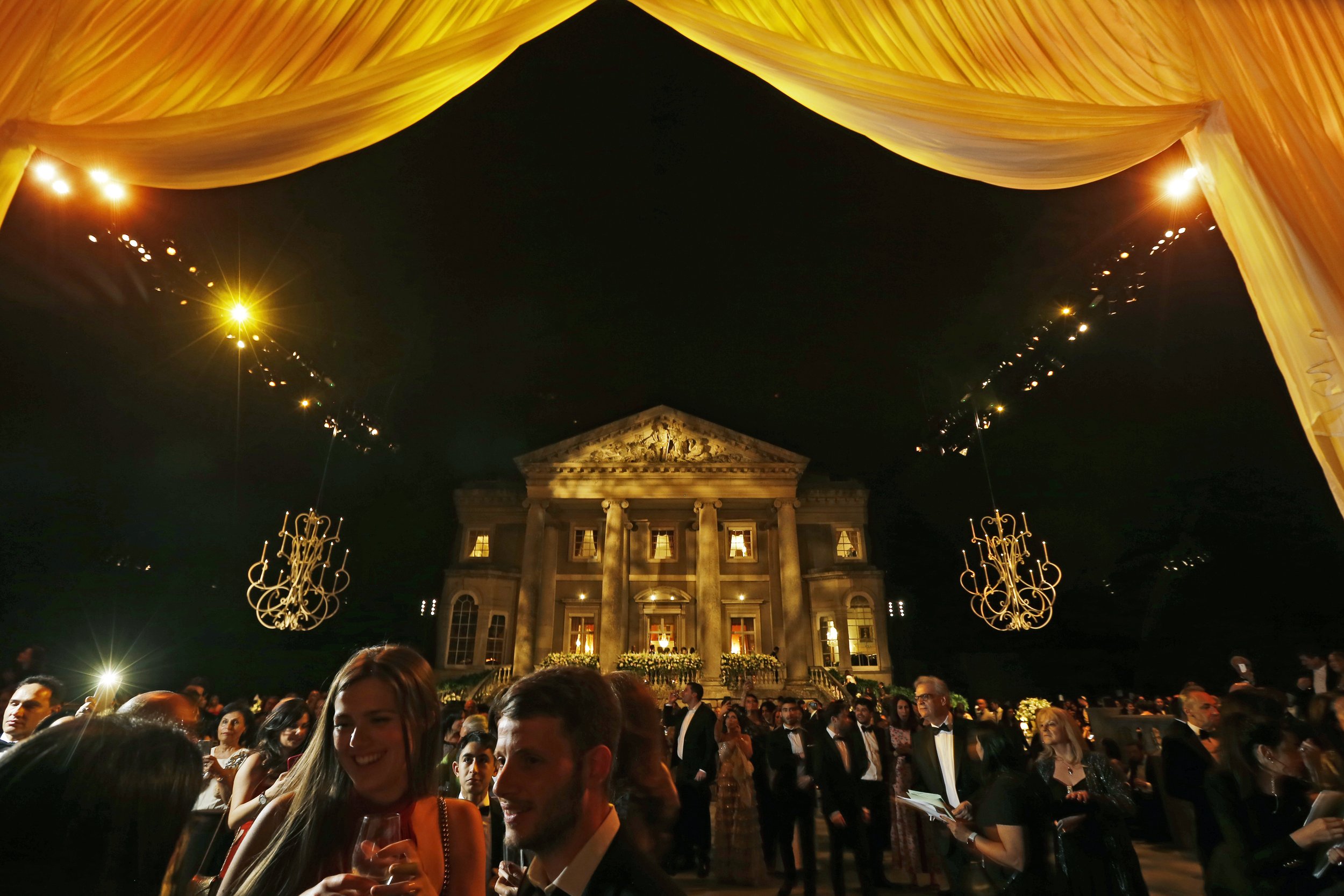 Nighttime elegant outdoor party with a large, illuminated mansion in the background, surrounded by guests dressed in formal attire, chandeliers, and a yellow draped canopy above.