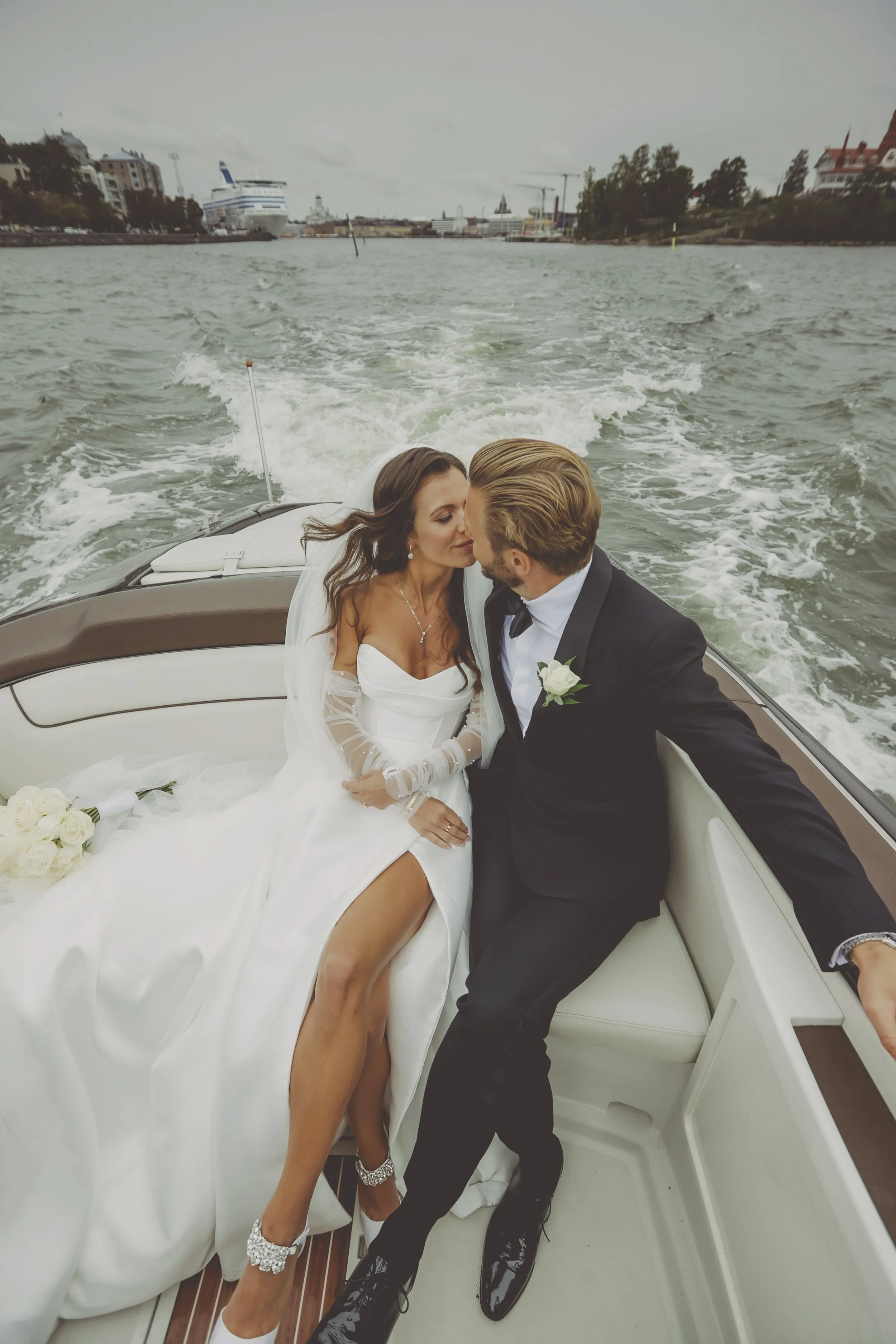 Bride and groom sharing a kiss on a boat, woman in white wedding dress, man in black suit, with a waterway and cityscape in the background.