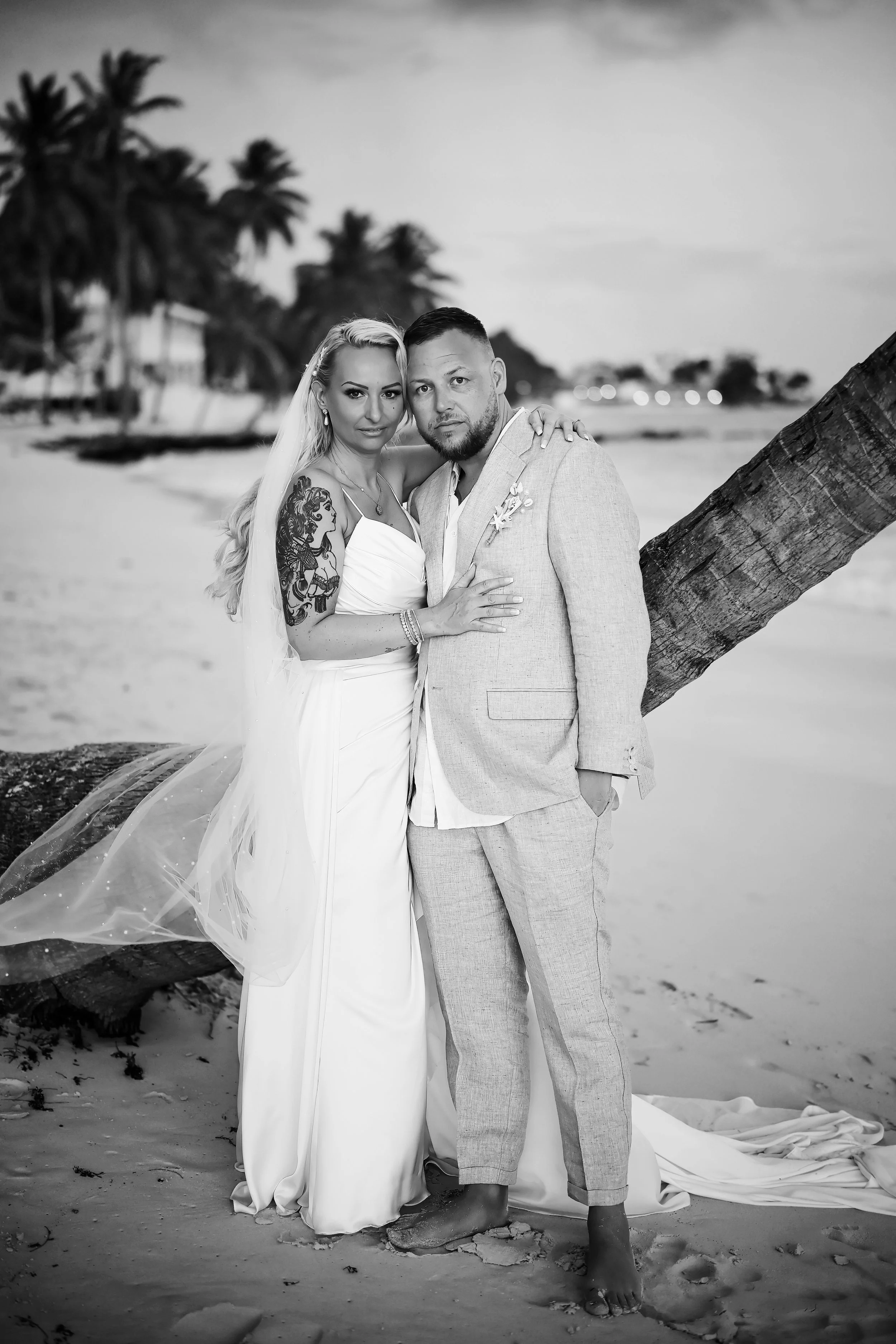 A black and white photo of a couple on a beach, standing close together with their arms around each other, near a leaning palm tree. The woman is wearing a white dress and has a tattoo on her arm, while the man is in a light-colored suit. There are t