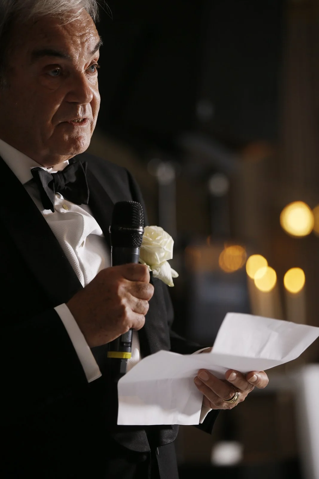 An elderly man in a tuxedo with a white rose boutonniere reading from a paper while holding a microphone.