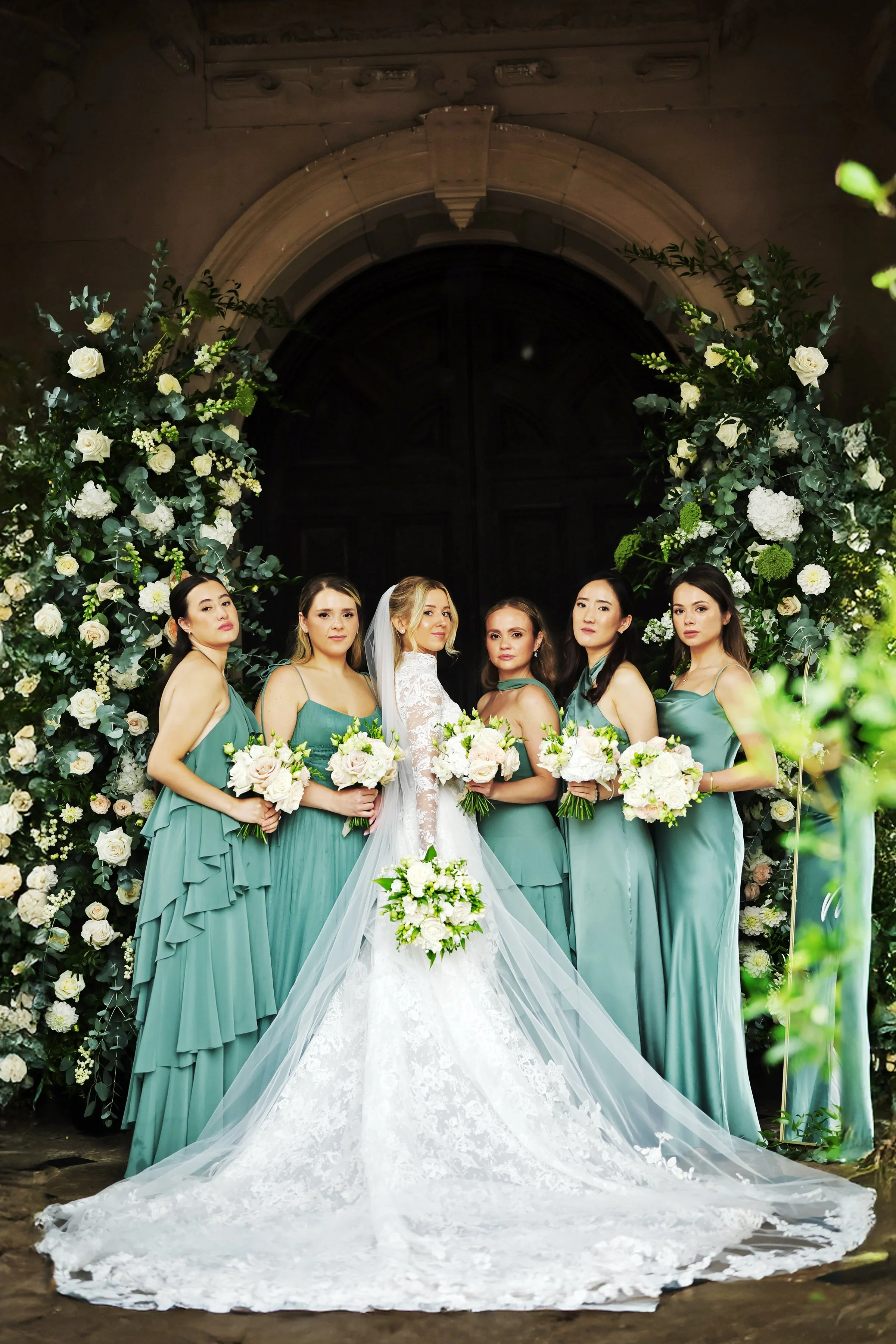 A bride in a white wedding gown with lace details and a long train, standing with six bridesmaids in teal dresses, holding bouquets of white flowers, in front of a floral arch at a wedding ceremony.