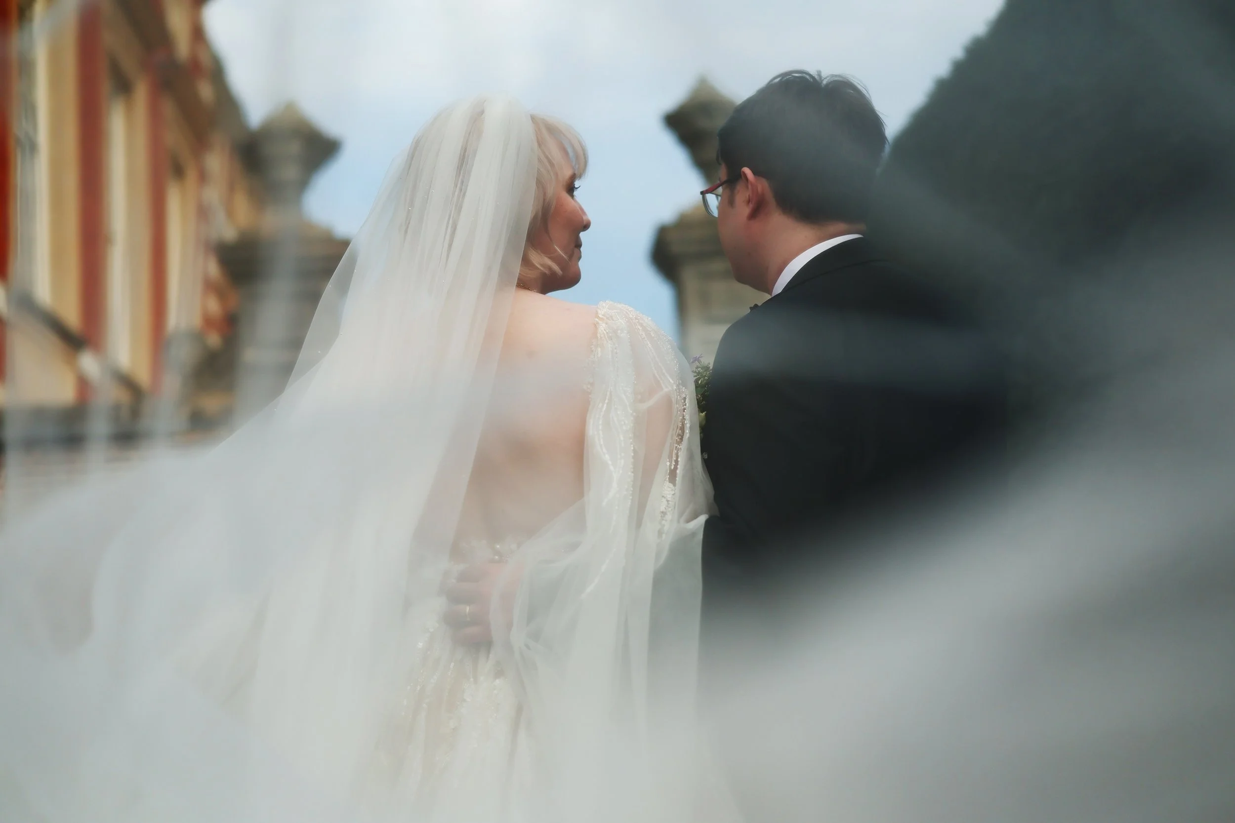 A bride and groom during their wedding ceremony in front of a historic building with ornate architecture, with the bride wearing a white wedding dress and veil and the groom in a black tuxedo, sharing a moment of intimacy.