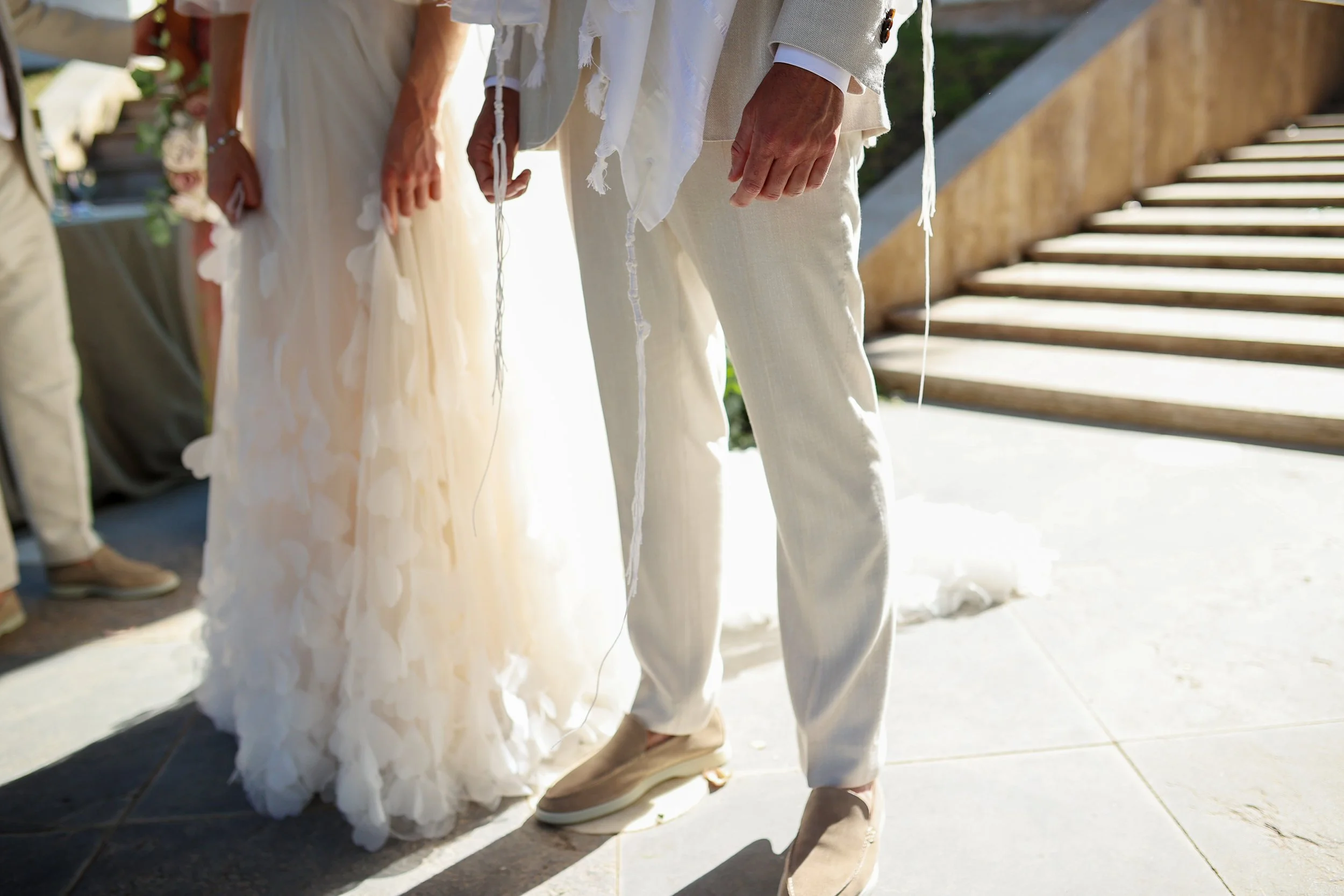 A couple standing outdoors, dressed in white and beige wedding attire, likely during a wedding ceremony. The focus is on their hands and lower bodies, with a staircase and green surroundings in the background.