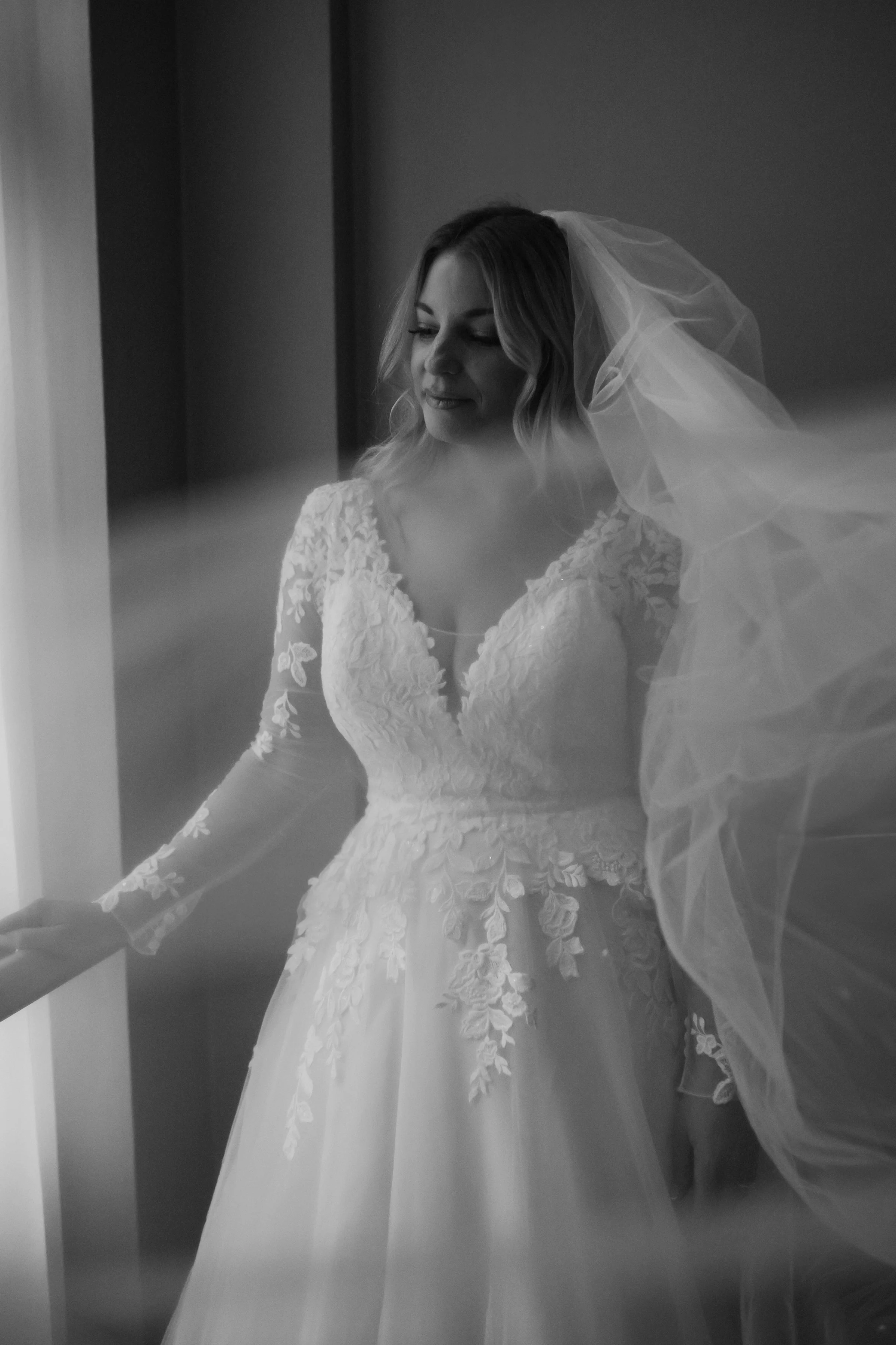 A woman in a wedding dress with lace details and a veil, standing near a wall and looking down with a gentle expression.
