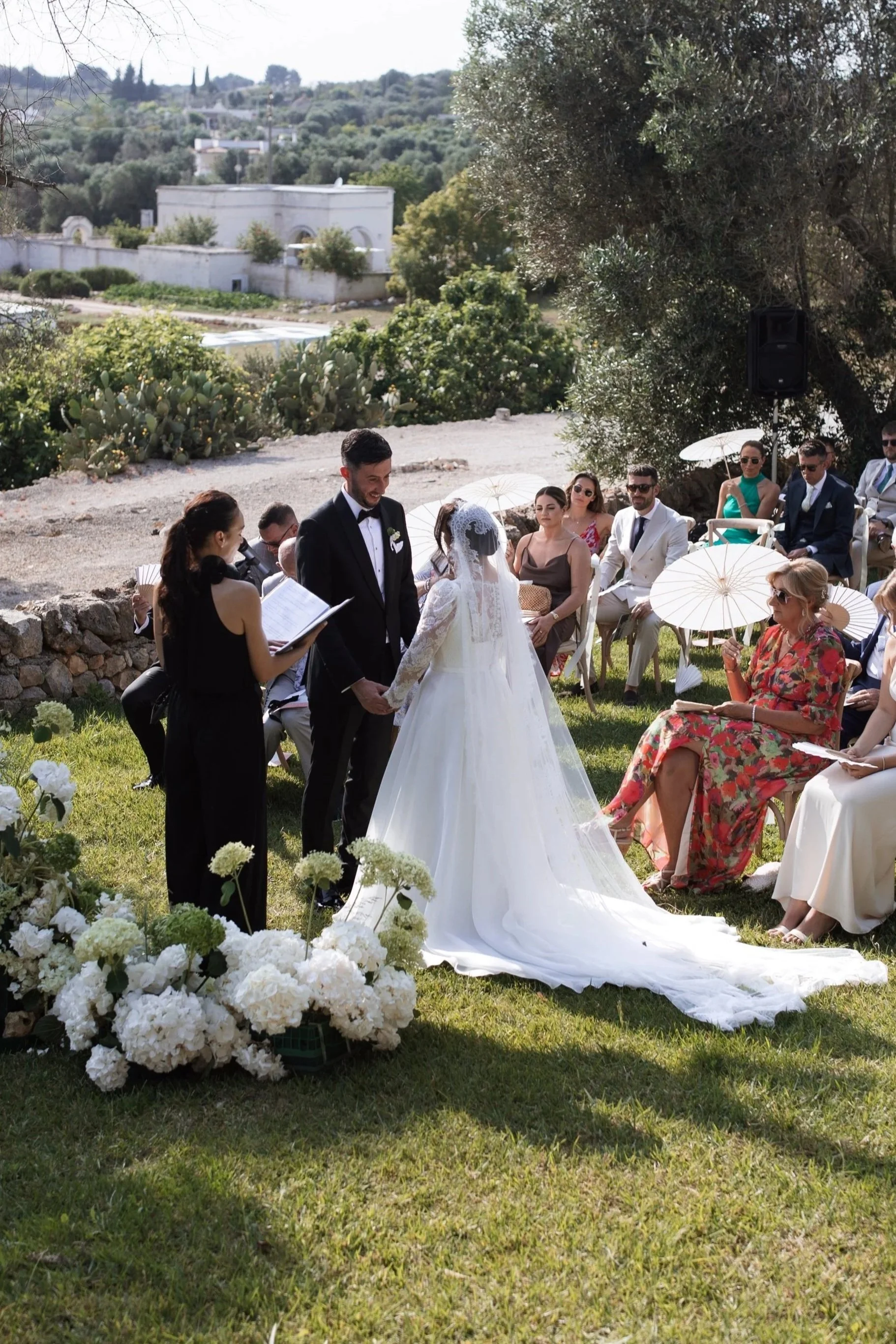 A wedding ceremony outdoors with the bride and groom holding hands. The bride is in a white wedding dress, and the groom in a black tuxedo. Guests are seated on chairs, some with umbrellas, under a tree. Flowers decorate the scene, and a person offic