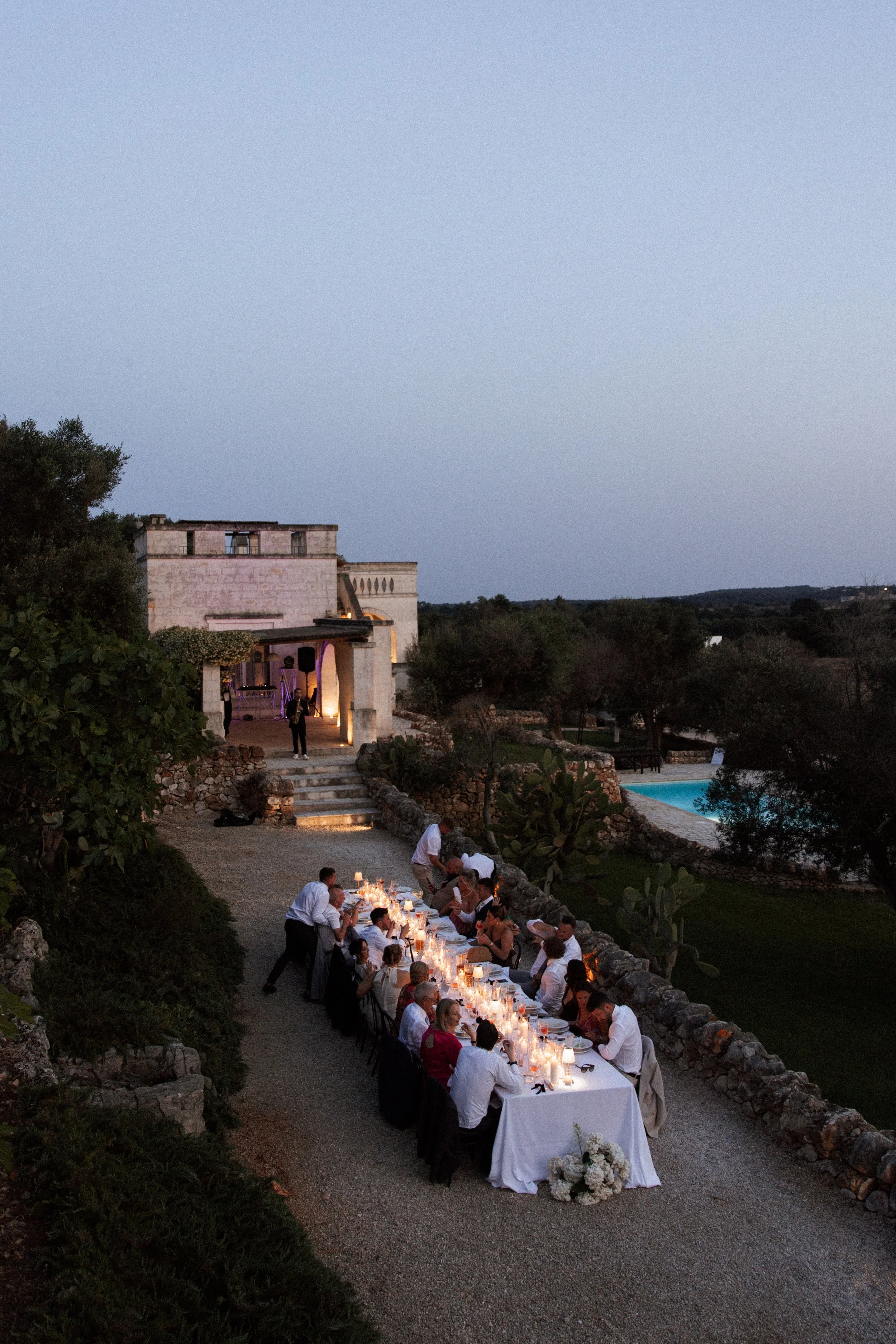 An outdoor evening dinner event with a long table set with candles and flowers, where guests are seated and servers are serving dinner, with a large building and trees in the background under a clear sky.