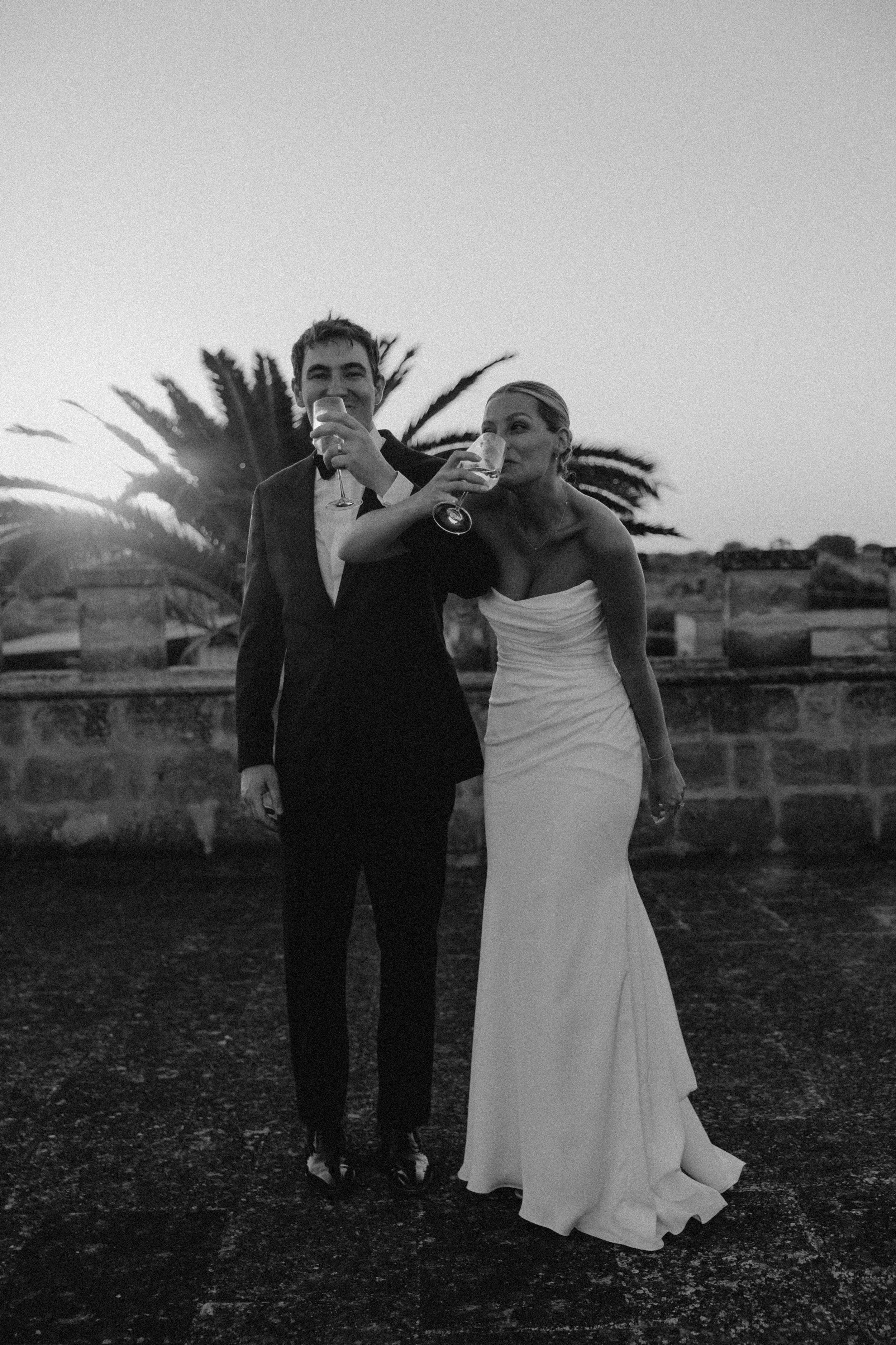 Black and white photo of a bride and groom standing outdoors, both holding glasses of champagne and smiling. The bride is wearing a strapless wedding gown and the groom is in a tuxedo. A large palm tree and low walls are visible in the background under a clear sky.