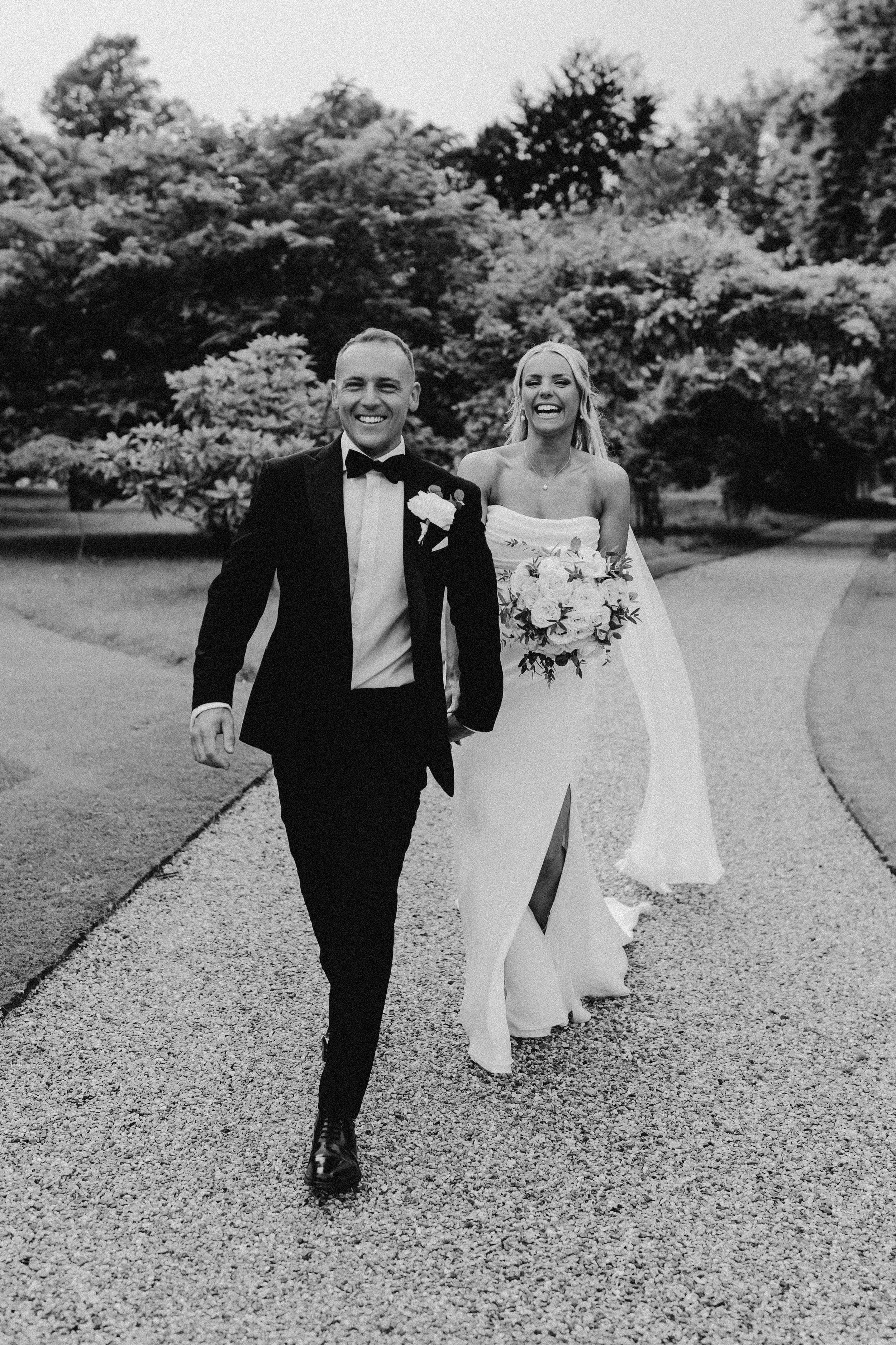 A black and white photo of a joyful bride and groom walking on a gravel path with trees in the background. The groom is wearing a tuxedo and the bride is in a strapless wedding dress holding a bouquet.