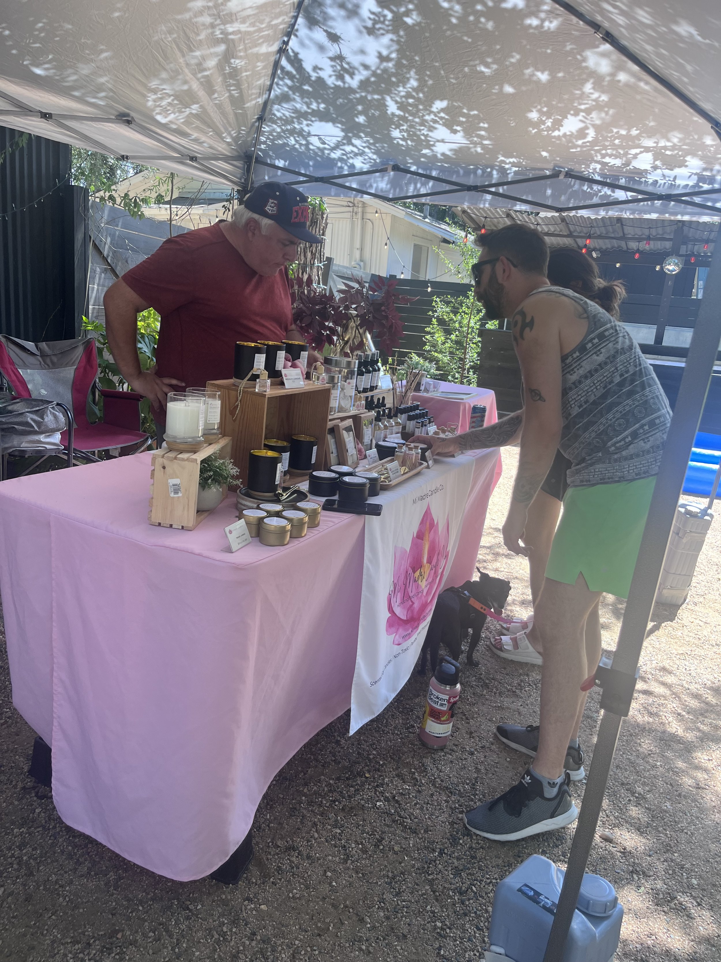 A man and a woman are at an outdoor market or craft fair under a canopy. The man, wearing a sleeveless shirt, shorts, and sneakers, is pointing at items on a table covered with pink fabric. The woman behind the table is wearing a red shirt and a base