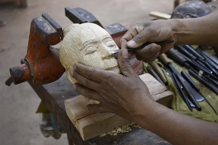 Close-up of a woodcarver's hands carefully carving a wooden face with traditional hand tools, showing detailed craftsmanship and the natural wood grain.