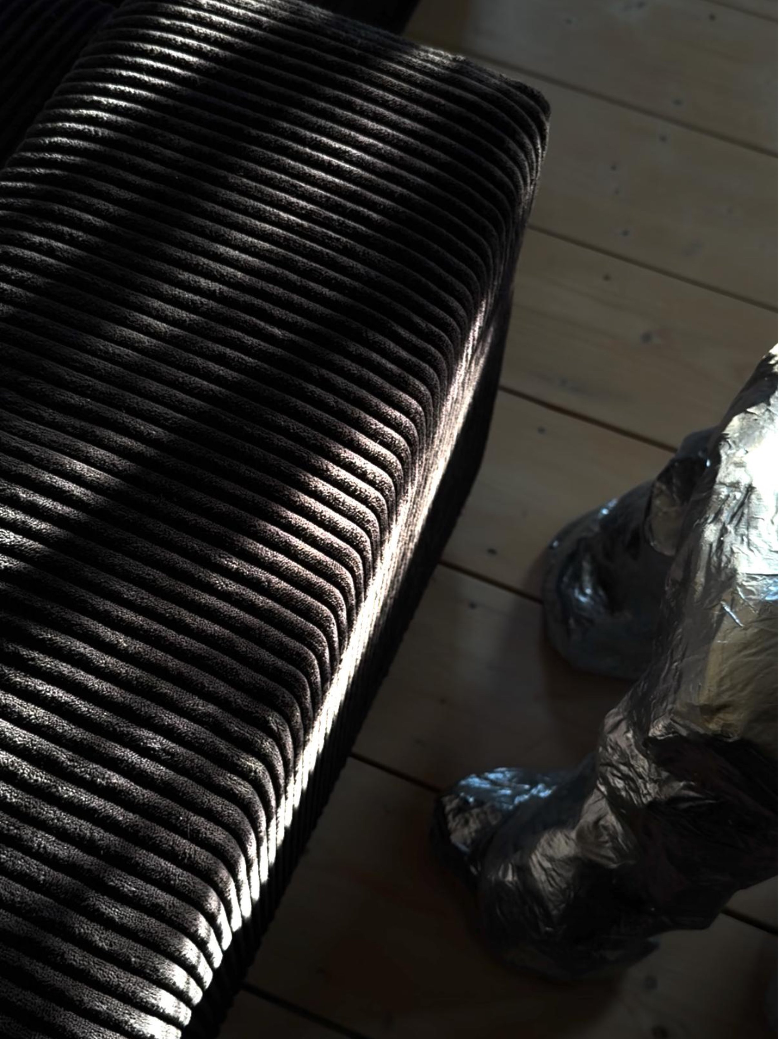 Nahaufnahme eines schwarzen, gerippten Gegenstands, vermutlich eine Matte, die an einem Fenster liegt, mit Sonnenlicht, das dass Muster beleuchtet, und ein Stück Aluminiumfolienverpackung auf Holzboden.