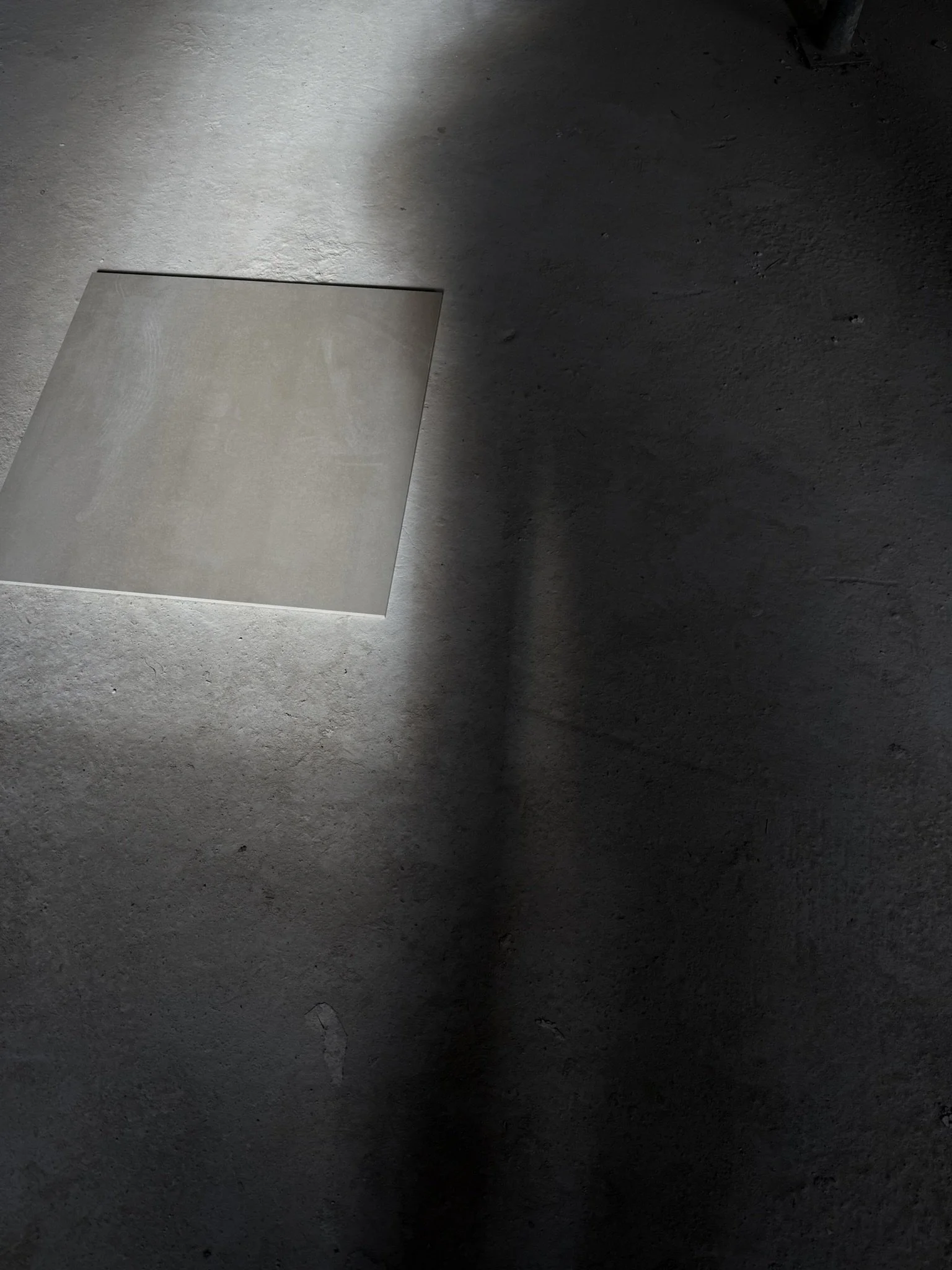 Eine dunkle, raue Betonwand mit einem quadratischen, hellen Metall-Panel, das an der Wand befestigt ist, und einem Schatten auf der Wand.