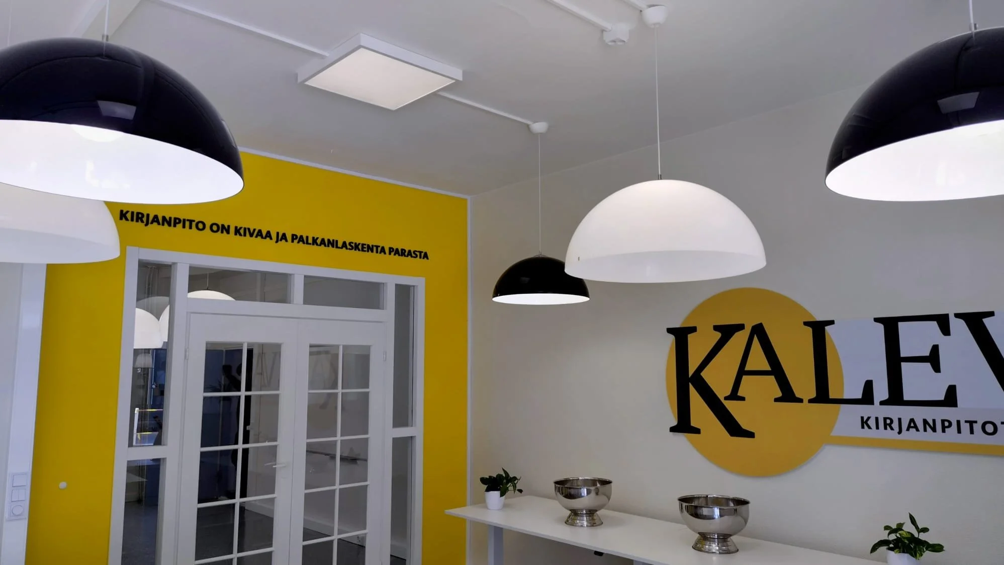 Sisustuskurssin tila, jossa on mustia ja valkoisia kattolamppuja, keltaisella taustalla oleva teksti 'KIRJANPITO ON KIVAA JA PALKANLASKENTA PARASTA', ja kookas K-A-L-E-kirjaimistin logo seinällä.