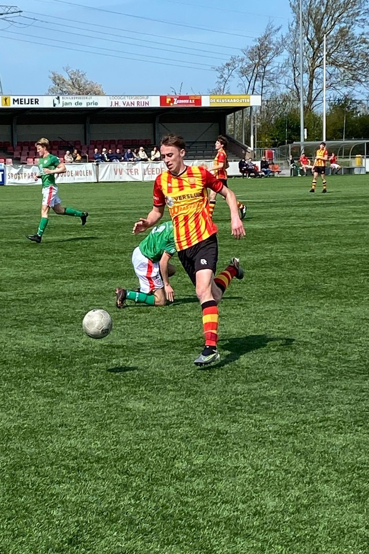 Voetballer in een rood-geel gestreept tenue die een bal trap op een grasveld, met andere spelers en toeschouwers op de achtergrond.