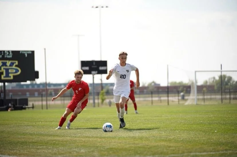 Twee jeugdvoetballers, een in een wit tenue met nummer 20 en een in een rood tenue, rennen achter een voetbal op een grasveld tijdens een wedstrijd.