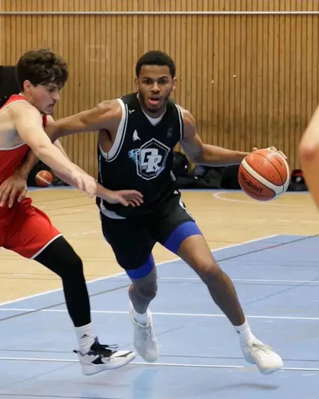 Twee basketballspelers in actie op een basketbalveld, één speler met een bal en de andere speler die probeert te verdedigen.