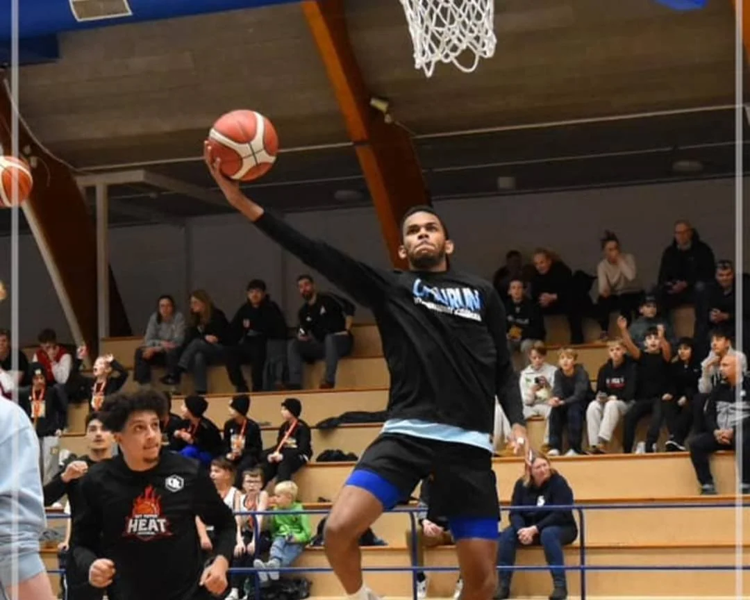 Man die basketball speelt in een indoor sporthal, met publiek op de tribune op de achtergrond.